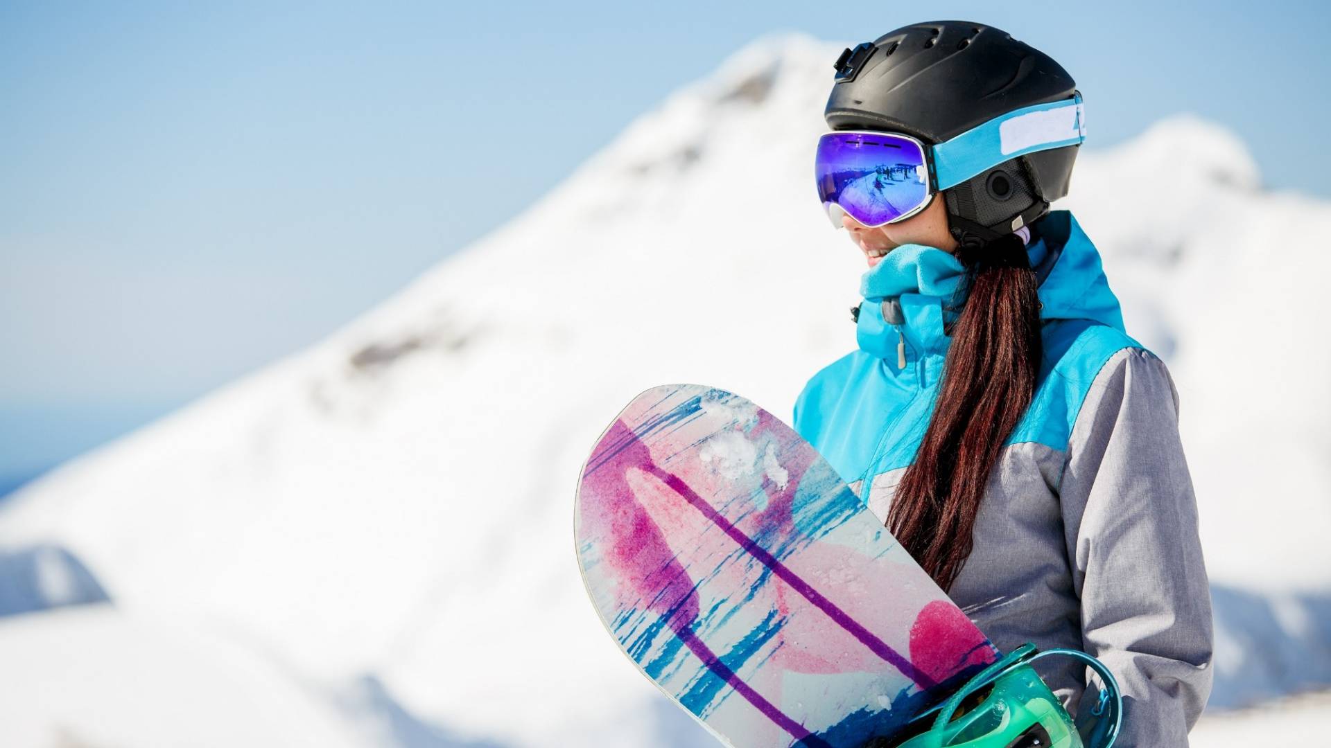 Photo of young woman tourist in helmet looking away with snowboard in hands against background of snow mountains