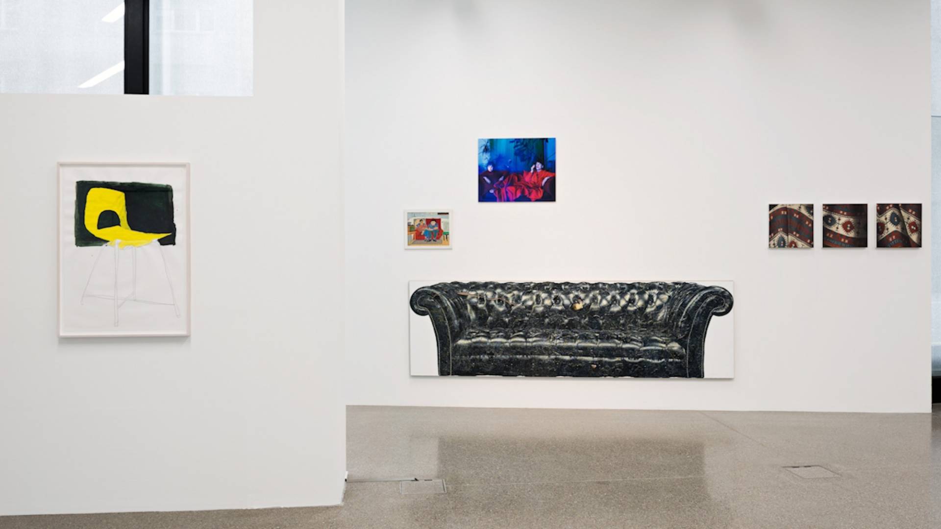 Blick in den Ausstellungsraum mit diversen Werken, die das Thema Sitzgelegenheiten darstellen: ein gelber Stuhl, ein schwarzes Sofa, sitzende Männer beim Radiohören, zwei Frauen auf einem Sofa und eine Darstellung Sigmund Freuds Sofa-Decke.