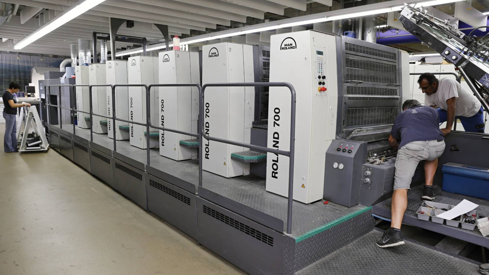 Eine Reihe von modernen Druckmaschinen. Zwei Männer arbeiten an einer der Maschinen.