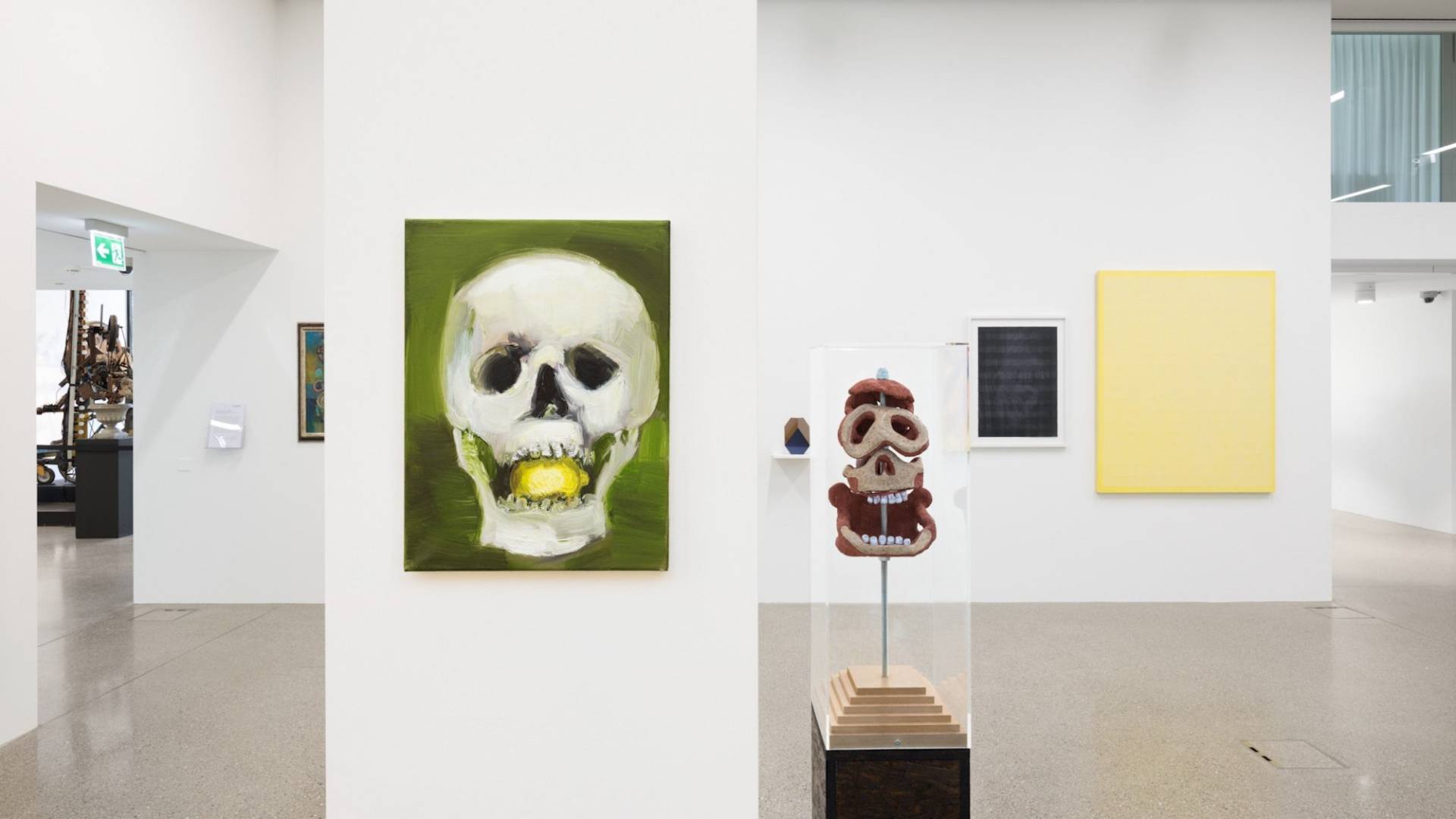 Im Vordergrund das Bild eines Totenkopfs (weiss auf grünem Grund) mit Zitrone im Mund und rechts davon eine Totenkopfskulptur aus Ton. 