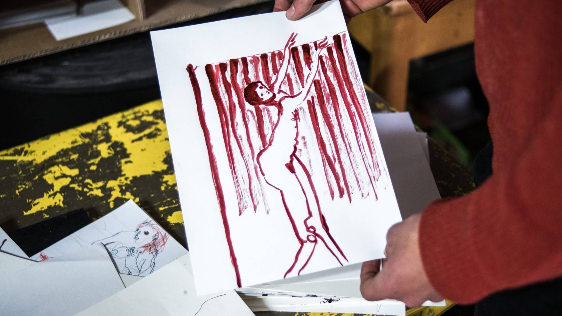 Andriu Deplazes hält eine Skizze in die Kamera – sie zeigt eine mit roter Tusche gezeichnete, tanzende Frau vor einem vorhangähnlichen Hintergrund. Im Hintergrund sind weitere, auf einem Tisch liegende Skizzen erkennbar.