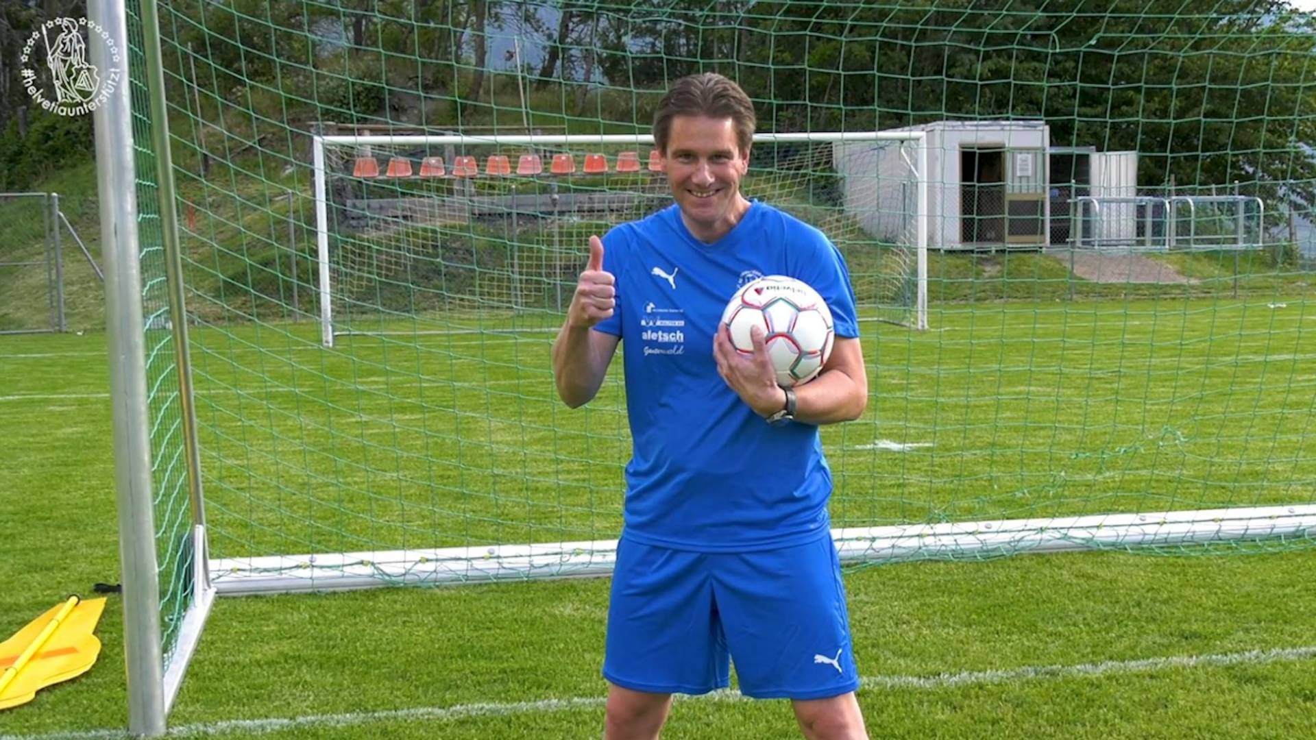A man smiles and gives a thumbs up in front of the goal.