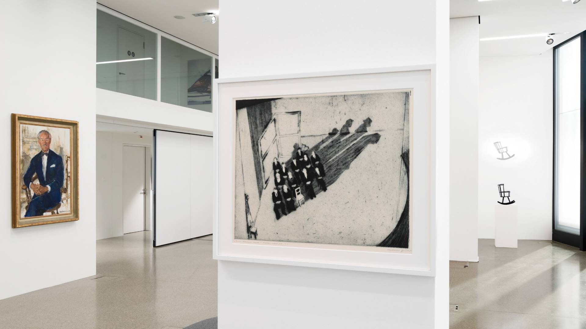 To the left hangs a luxuriously framed colour portrait of a man painted in oil. In the middle, a black-and-white etching entitled “Eine kleine Stadt”, to the right one of the installations with a small black wooden rocking chair.