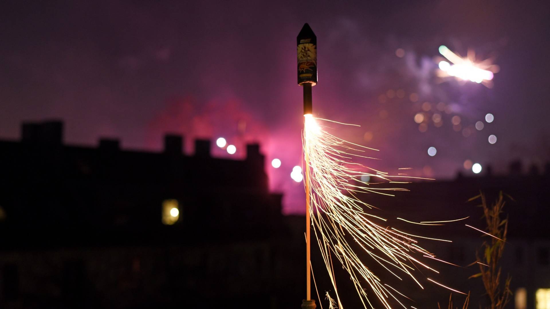 Sparks fly out of a firework rocket. In the background, we can see the night sky illuminated by fireworks.