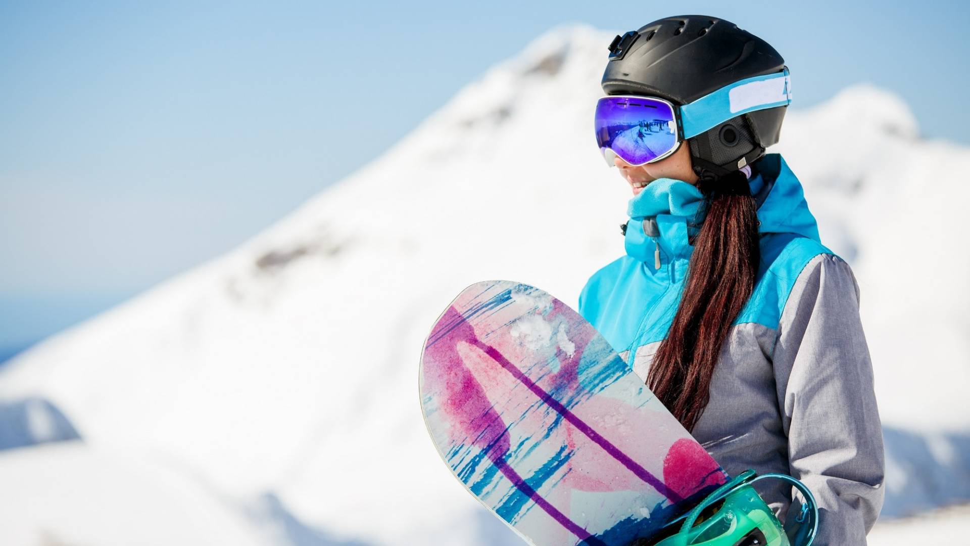 Photo of young woman tourist in helmet looking away with snowboard in hands against background of snow mountains