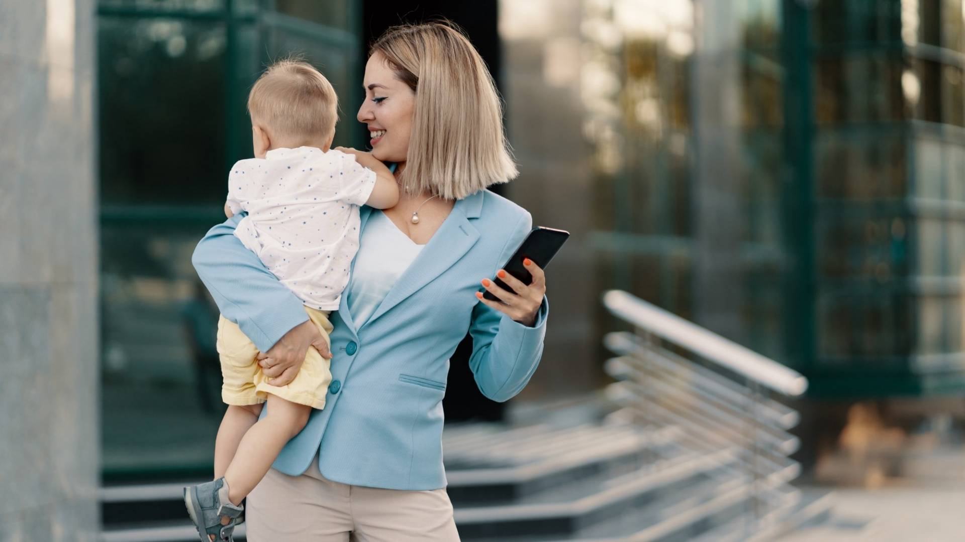 Successful business woman in blue suit with baby. Successful mother in business walking with baby in her hands and texting message on cellphone