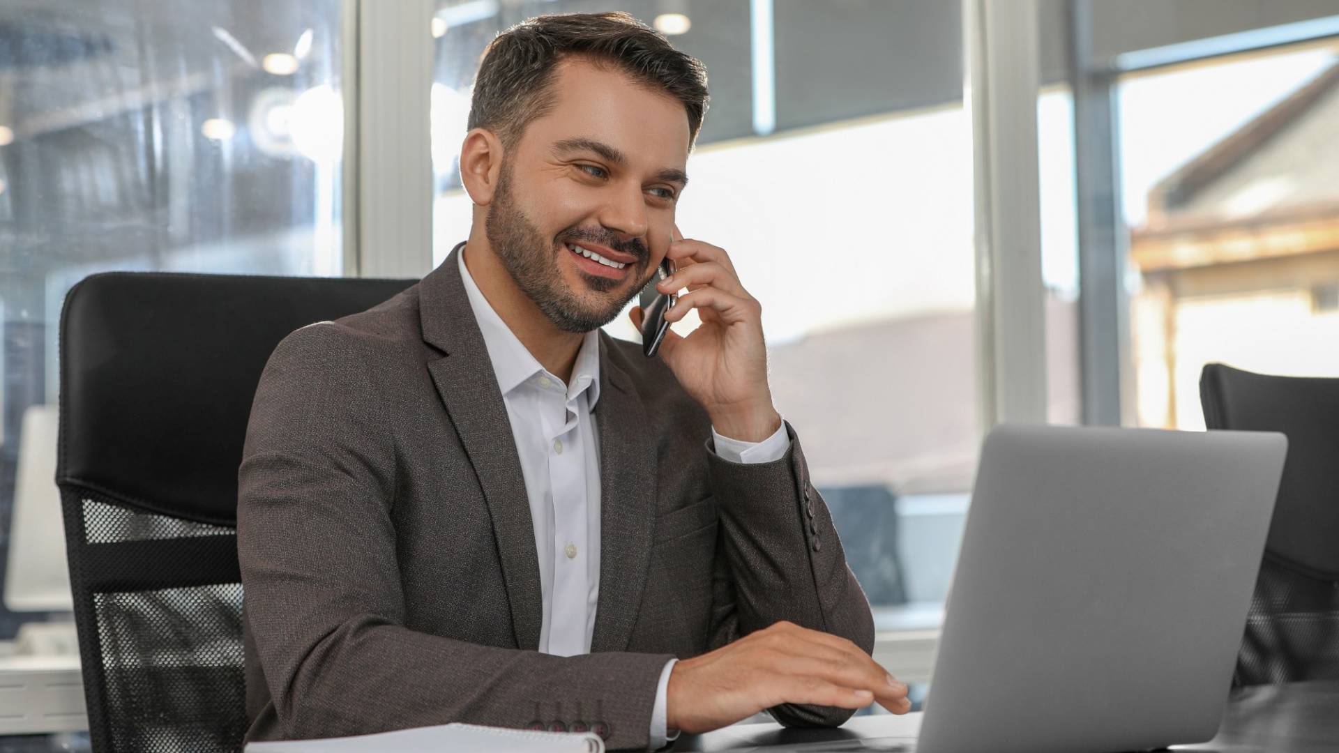 Happy man using modern laptop while talking on smartphone at black desk in office