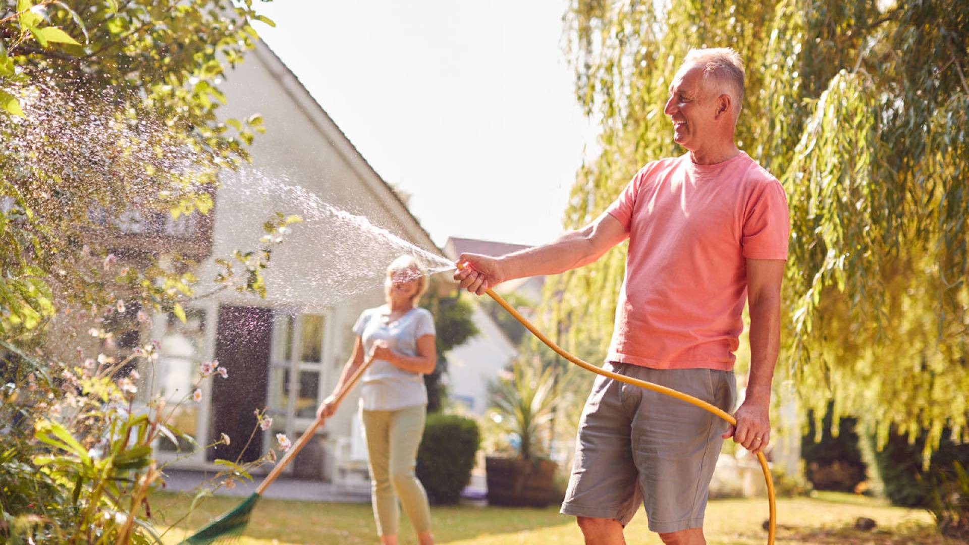 Retired Couple At Work Watering Plants With Hose And Tidying Garden With Rake At Home