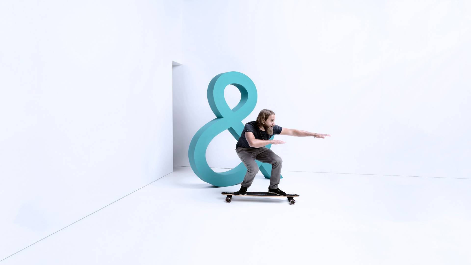 Un uomo si diverte visibilmente ad attraversare una stanza bianca su uno skateboard. 