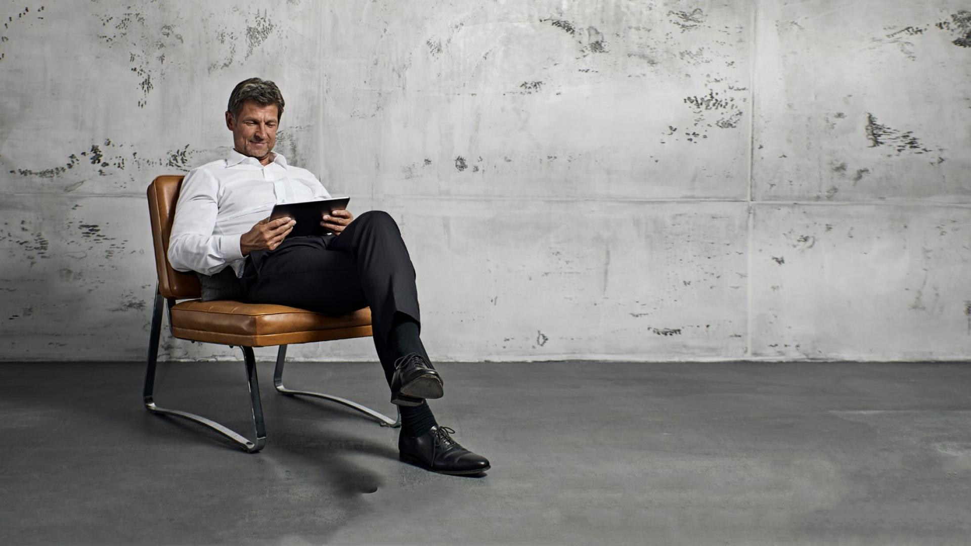 Ein erwachsener Mann mit weißem Hemd und schwarzer Anzughose sitzt auf einem Stuhl und hält ein Tablet in der Hand.