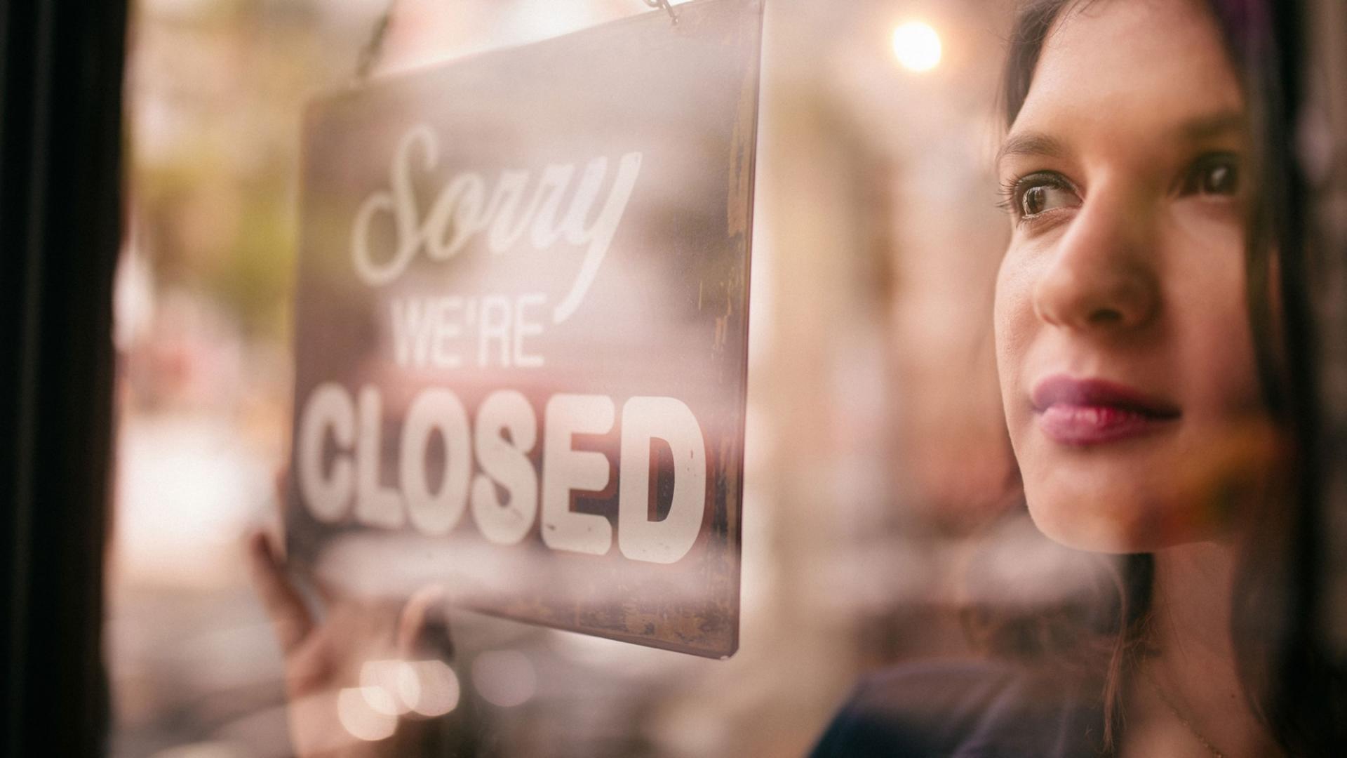 Frau ist hinter einer Glasscheibe, mit einem Schild auf dem "Sorry we´re closed" geschrieben ist, zu sehen.
