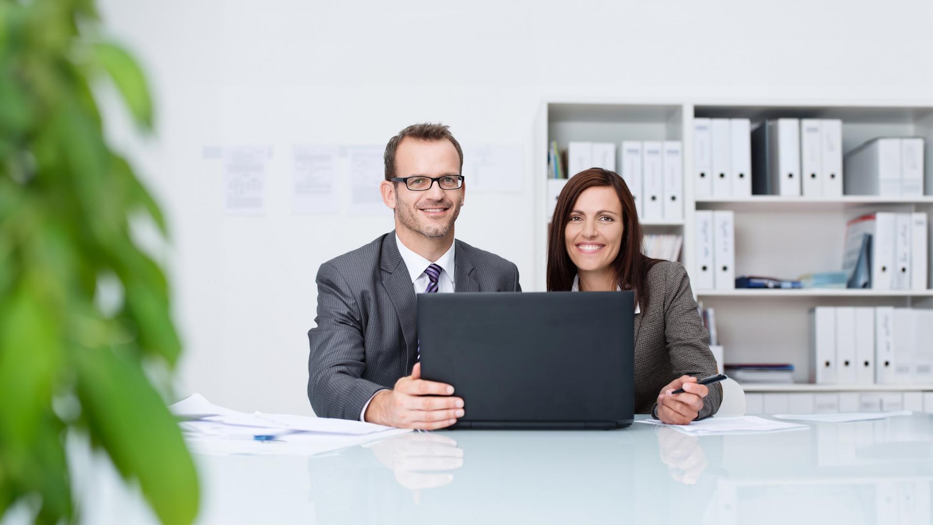 Ein Mann und eine Frau in Geschäftskleidung sitzen hinter einem Laptop und lächeln