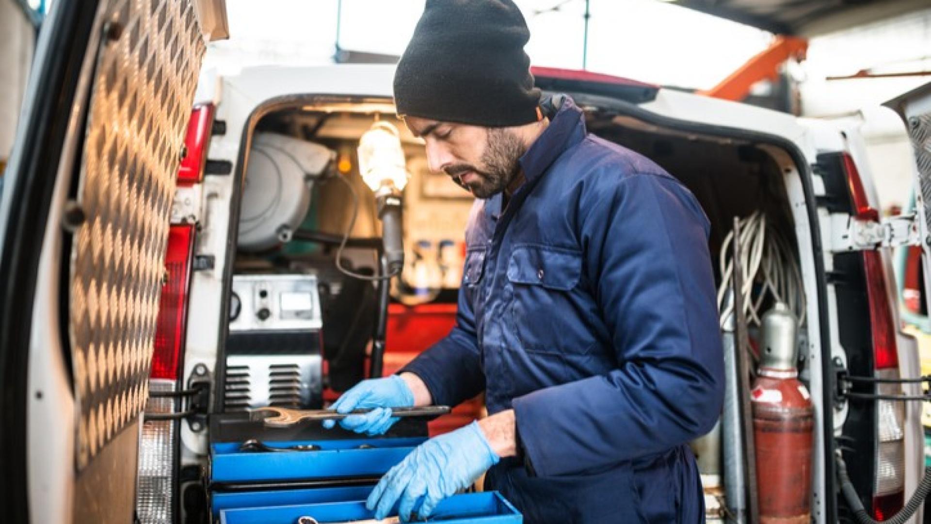 Ein Mann hält Werkzeug in der Hand und steht vor einem Transporter mit geöffnetem Kofferraum.