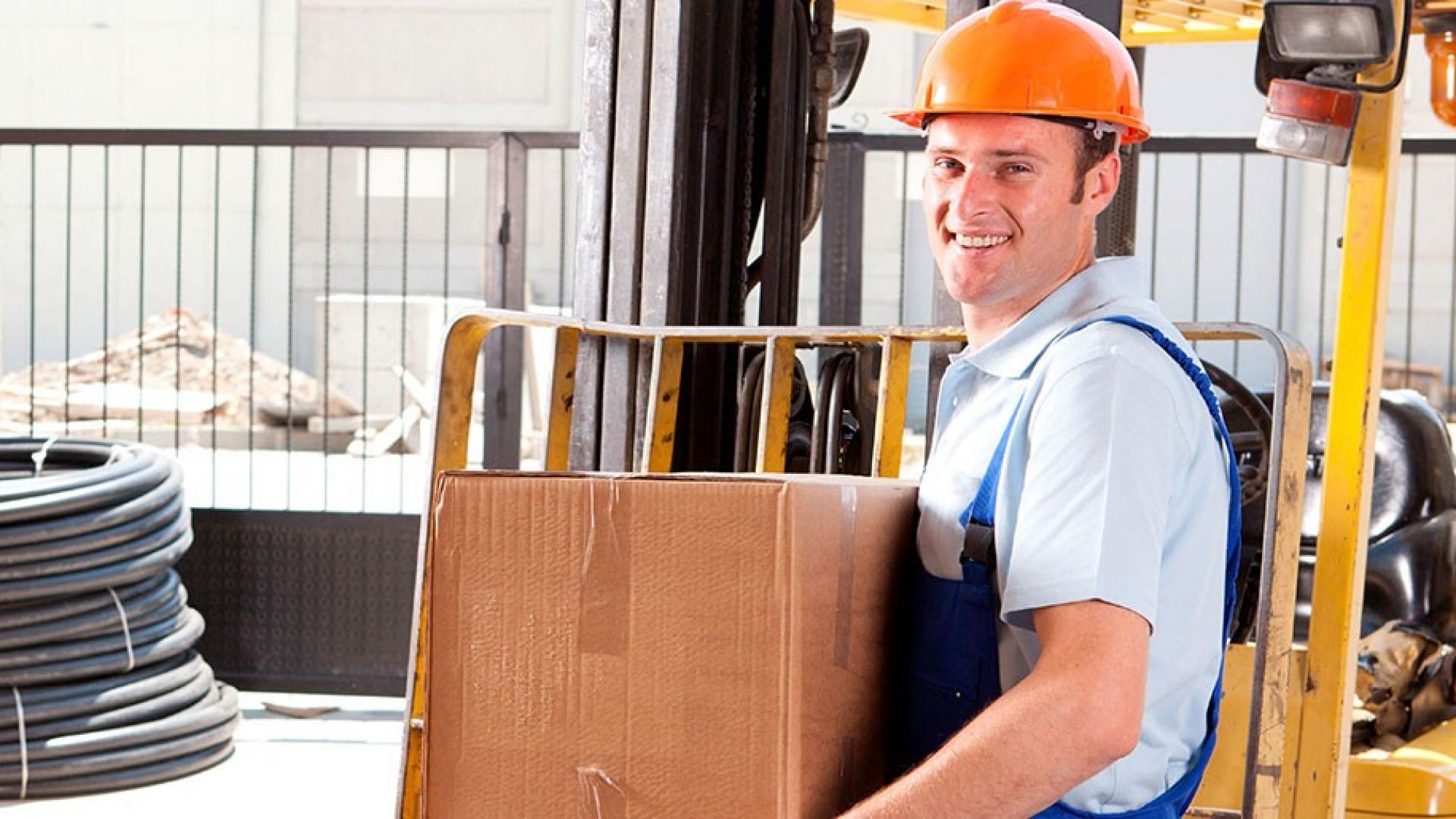 Ein Mann mit orangenem Helm und Blaumann hält einen Karton in der Hand und lächelt. 