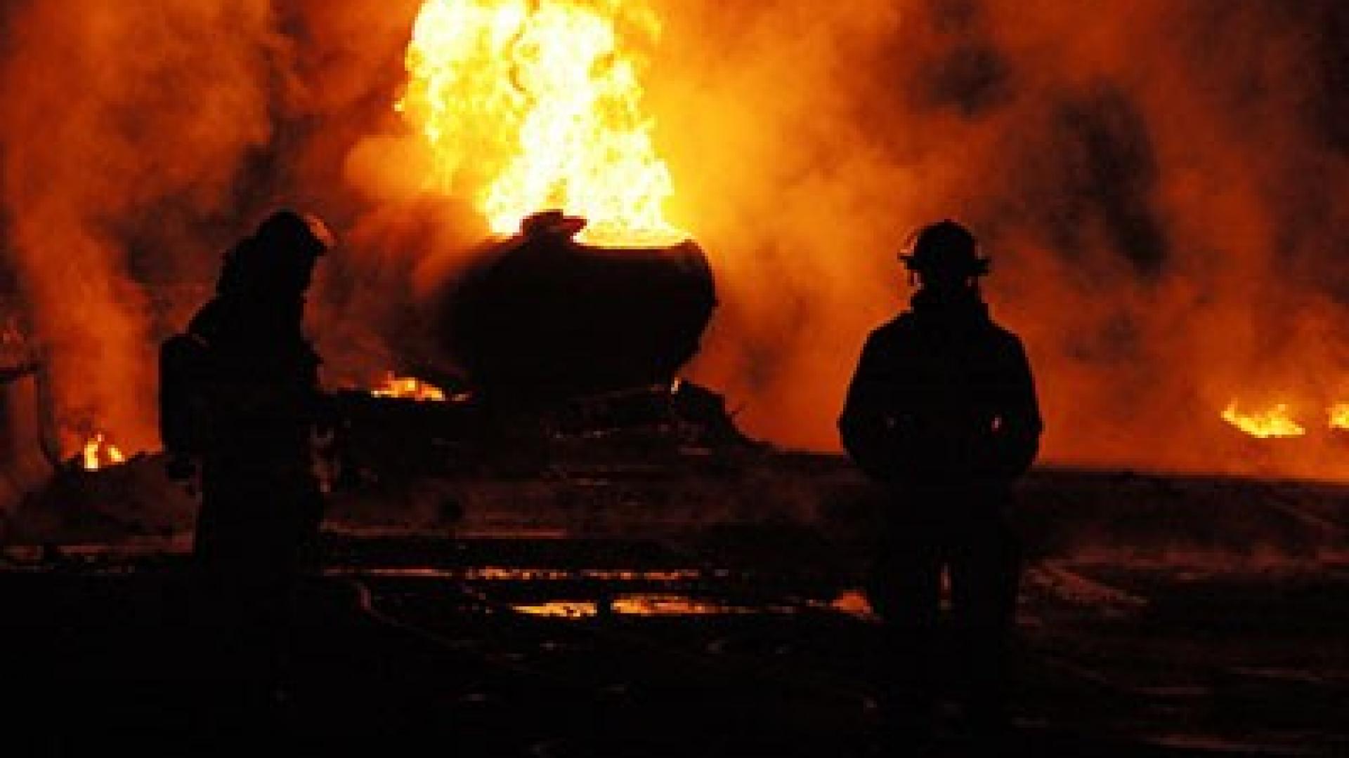 Ein brennendes Objekt vor dem zwei Feuerwehrleute stehen.