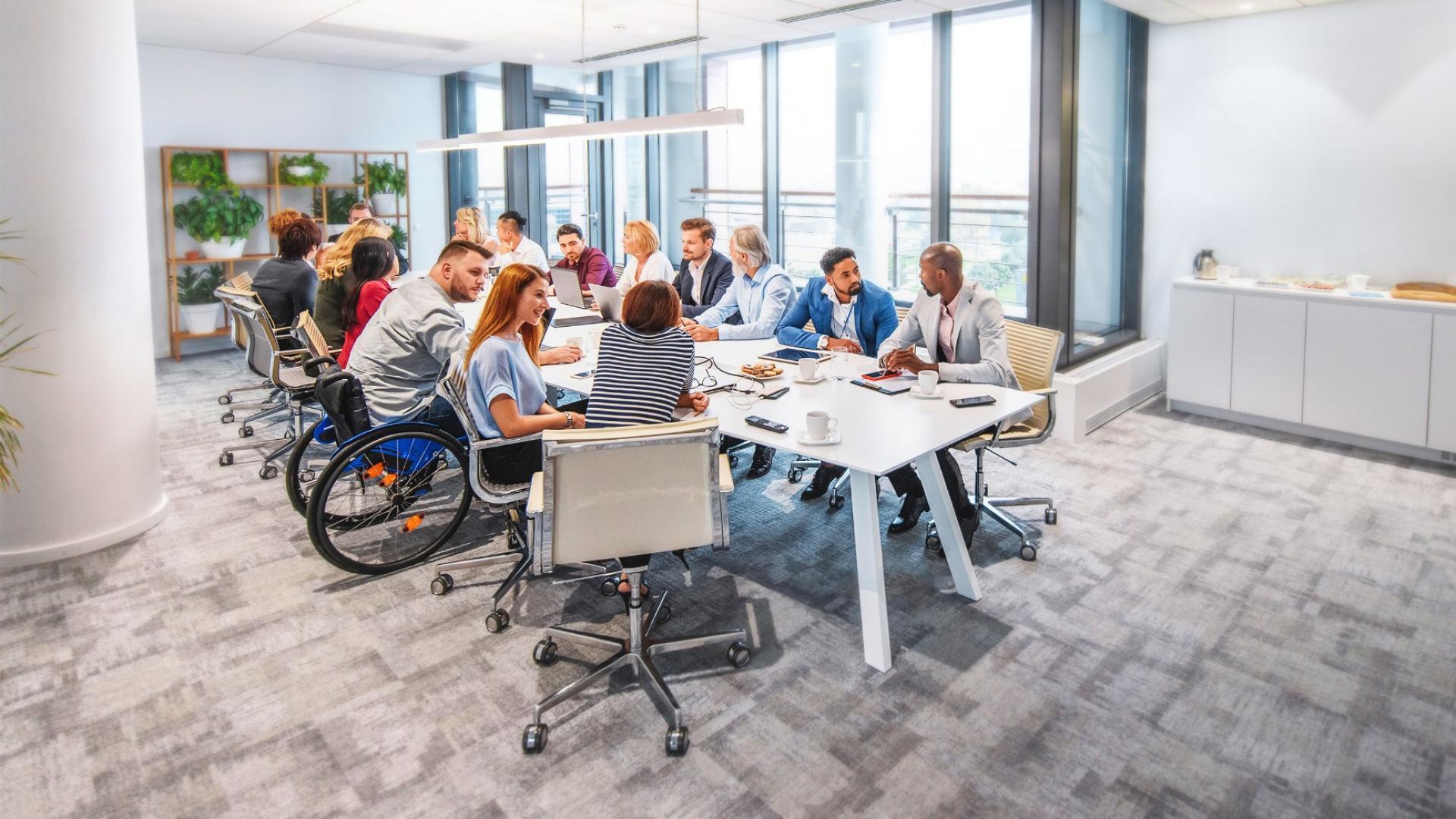 In einem Meetingraum sitzen mehre Personen, darunter auch ein Rollstuhlfahrer, an einme großen weißen Tisch.