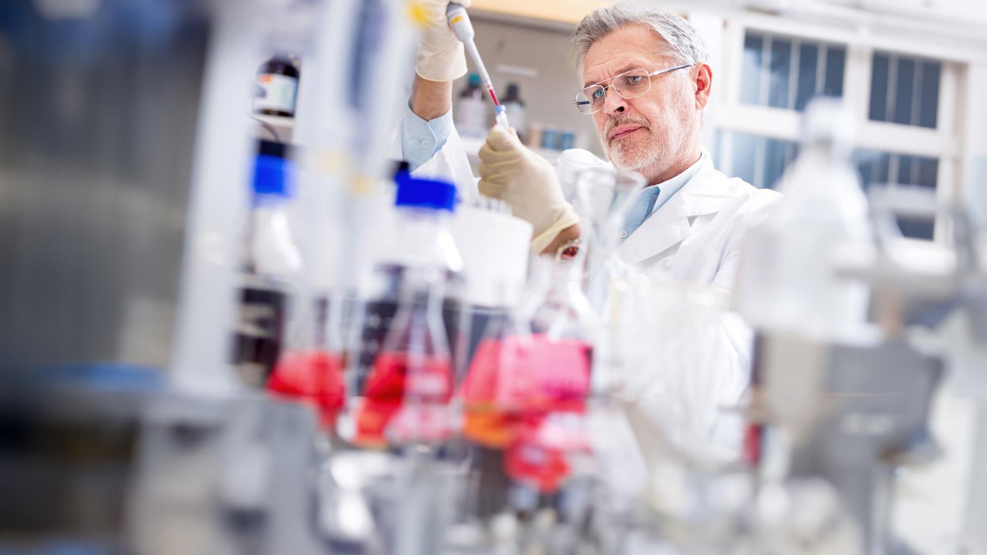 Ein Mann mit weißem Kittel hat eine Pipette in der Hand und füllt eine rote Flüssigkeit in eine Ampulle.