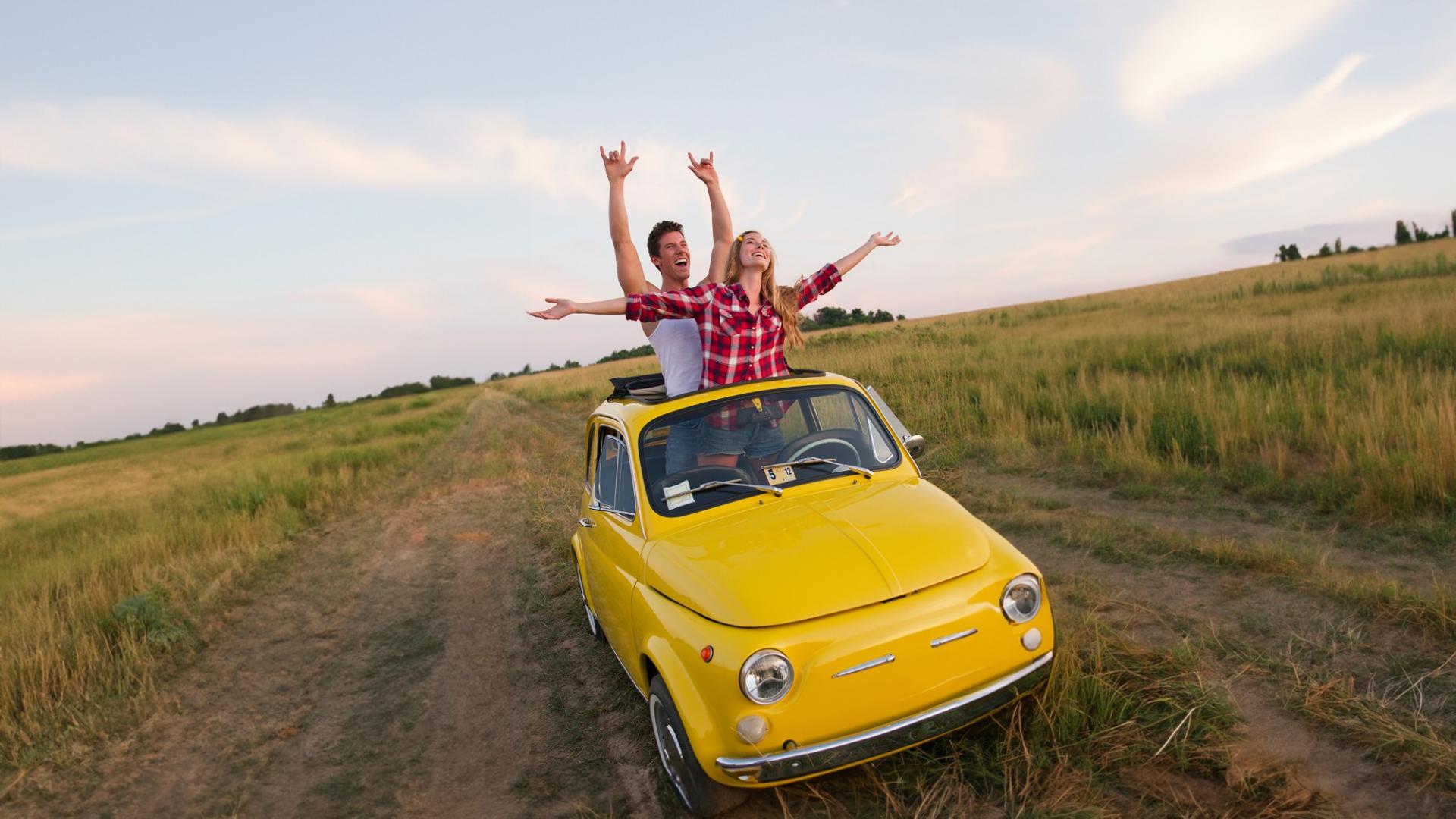Ein Mann und eine Frau stehen lächelnd in einem kleinen gelben Auto mit geöffneten Dach und breiten die Arme aus. 
