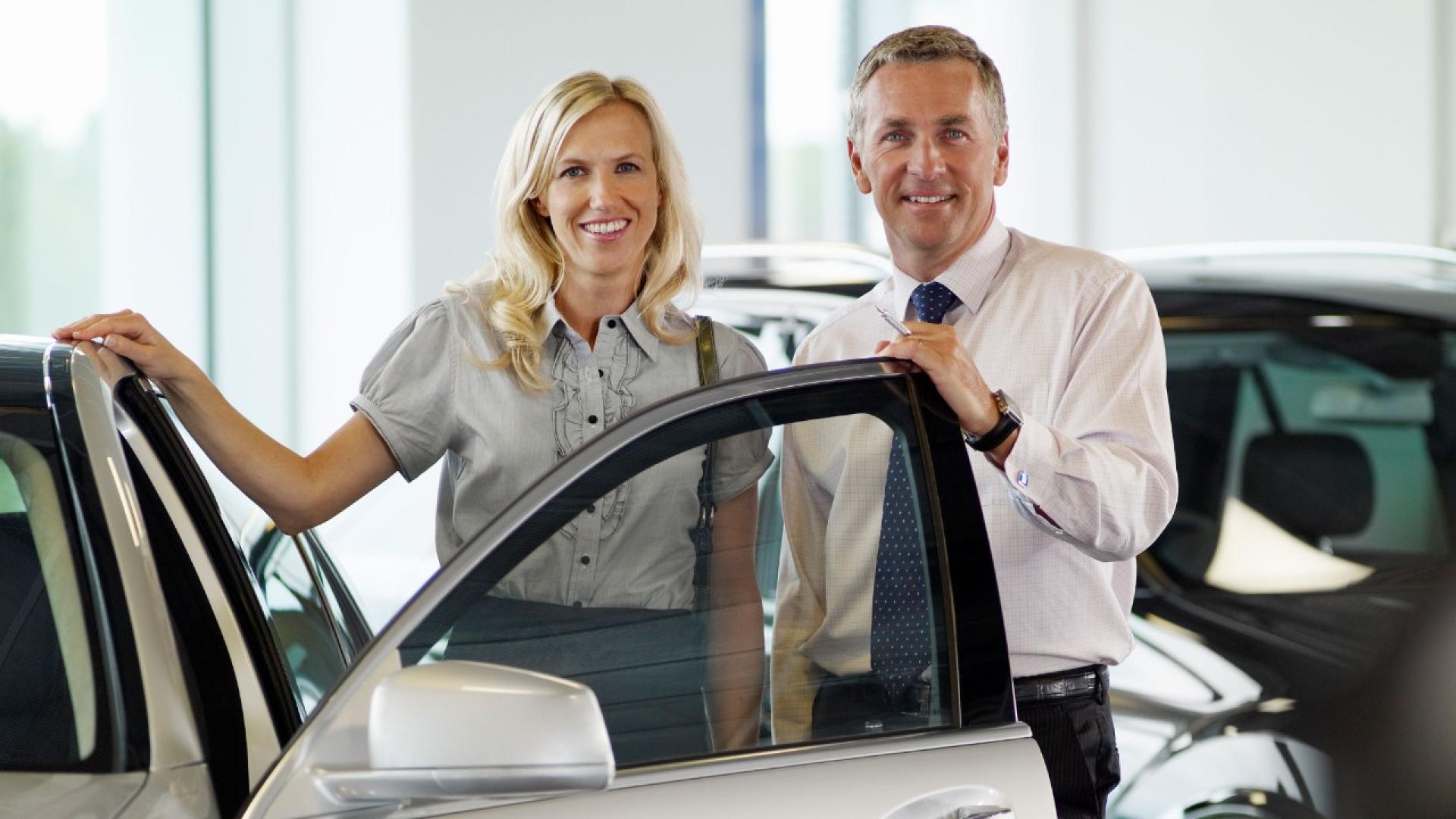 Ein Mann mit Hemd und Krawatte und eine Frau mit Bluse stehen lächelnd hinter einer geöffneten Autotür.