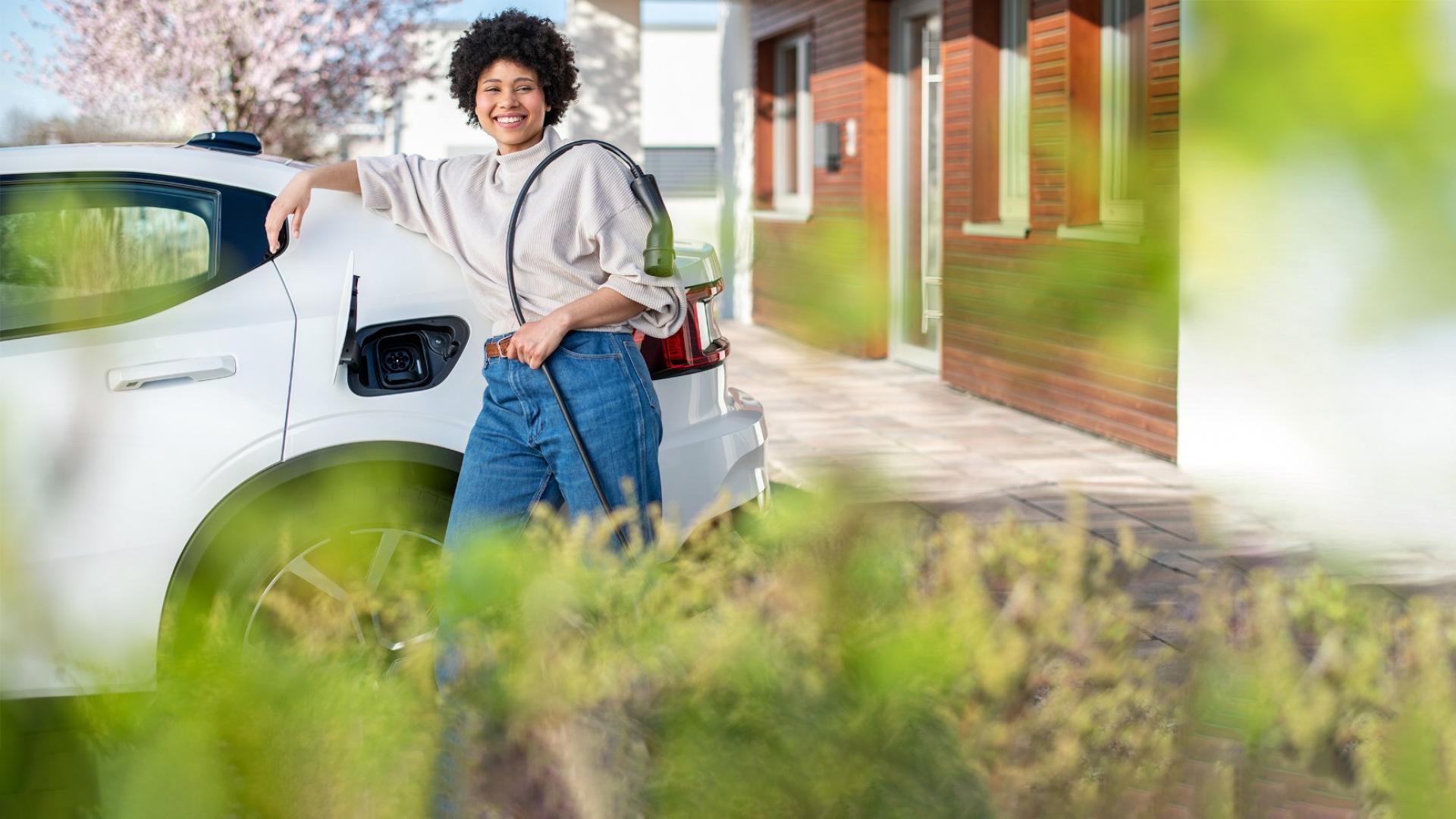 Auf dem Bild ist eine junge Frau vor ihrem E-Auto zu sehen, die gerade ein Ladekabel in der Hand hält, um ihr Elektroauto aufzuladen.