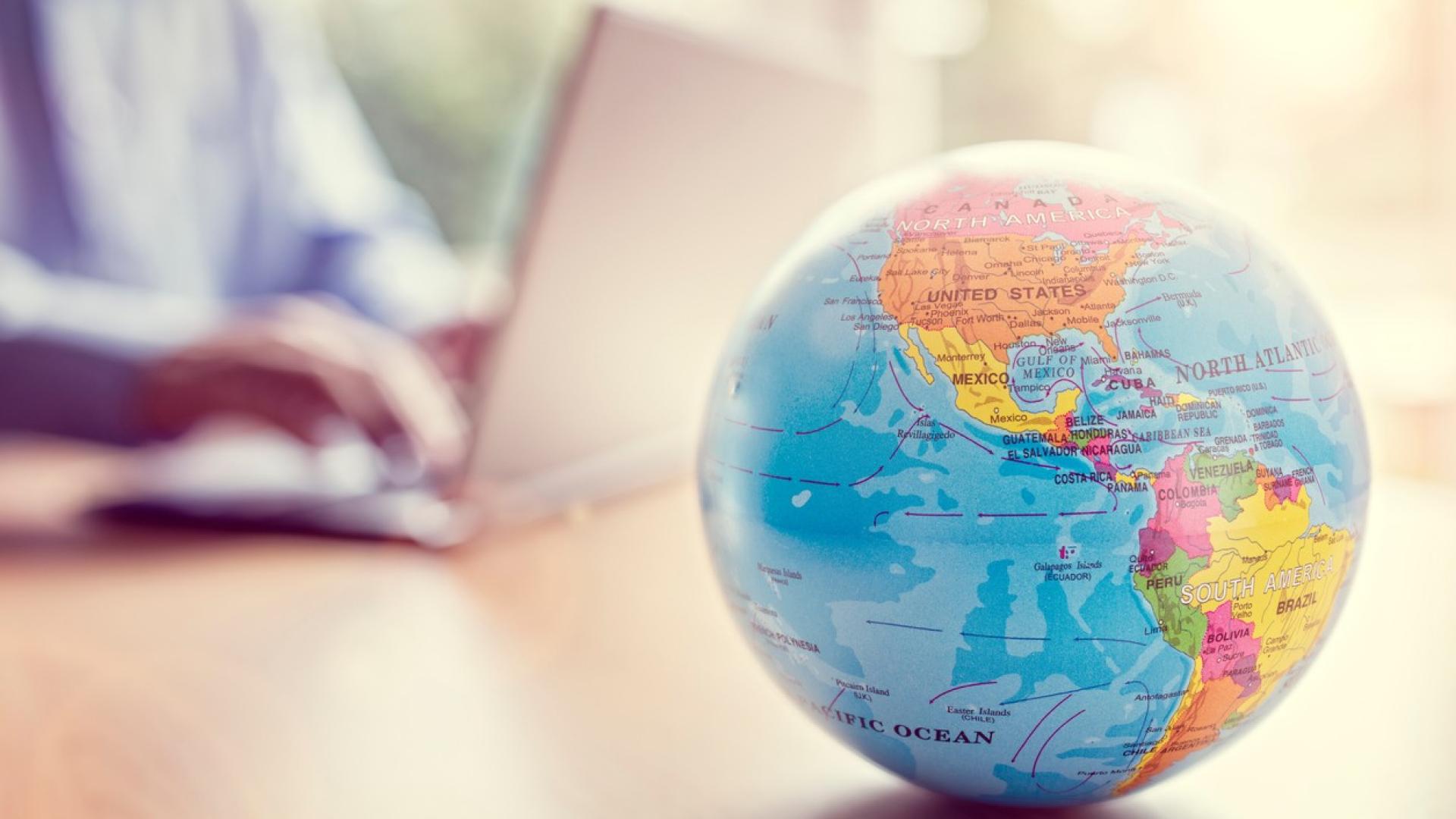 Weltkugel auf einem Schreibtisch – im Hintergrund sitzt eine Person am Laptop.
