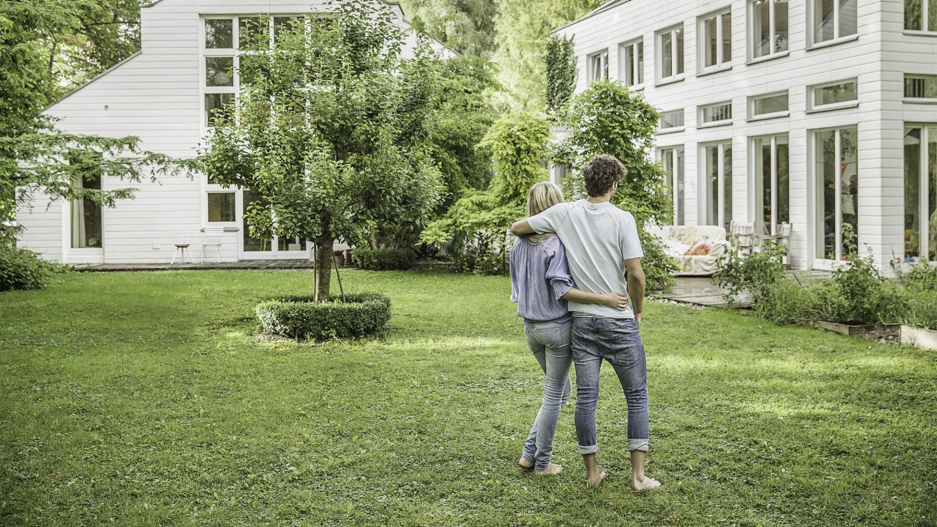 Ein Mann und eine Frau stehen eng umschlungen auf einer grünen Wiese und blicken auf ein Haus.