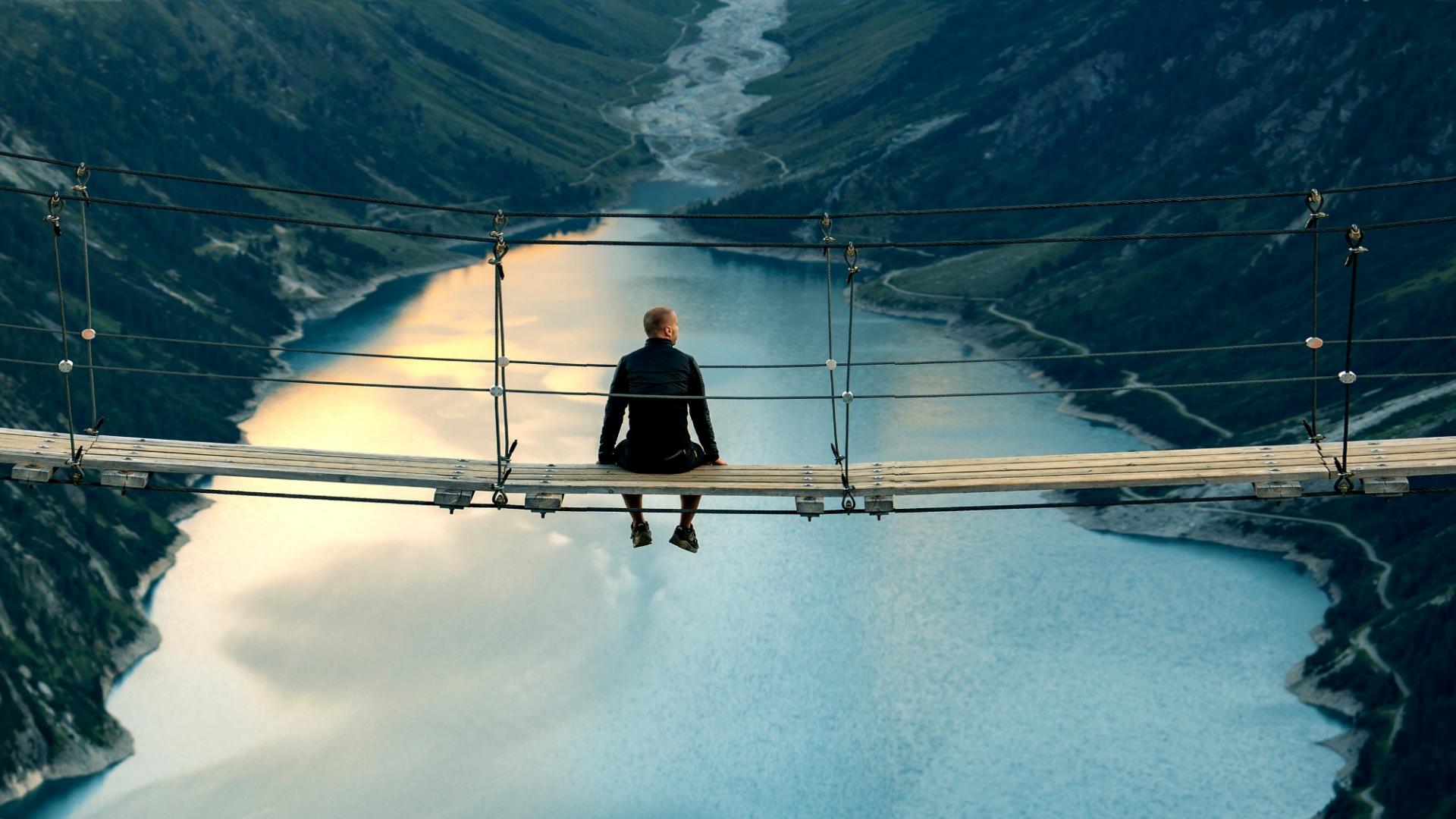 Ein Mann sitzt auf einer hohen Hängebrücke und blickt auf einen großen See mit Bergpanorama.