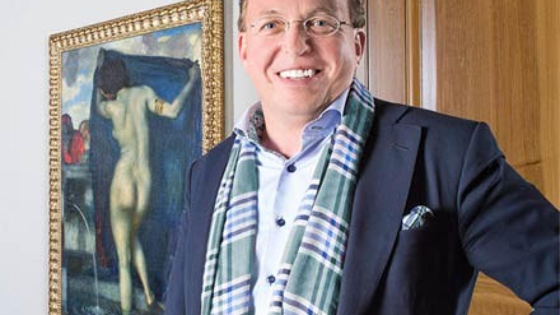 Ein erwachsener Mann mit Anzug und Brille steht vor einem Gemälde und lächelt.