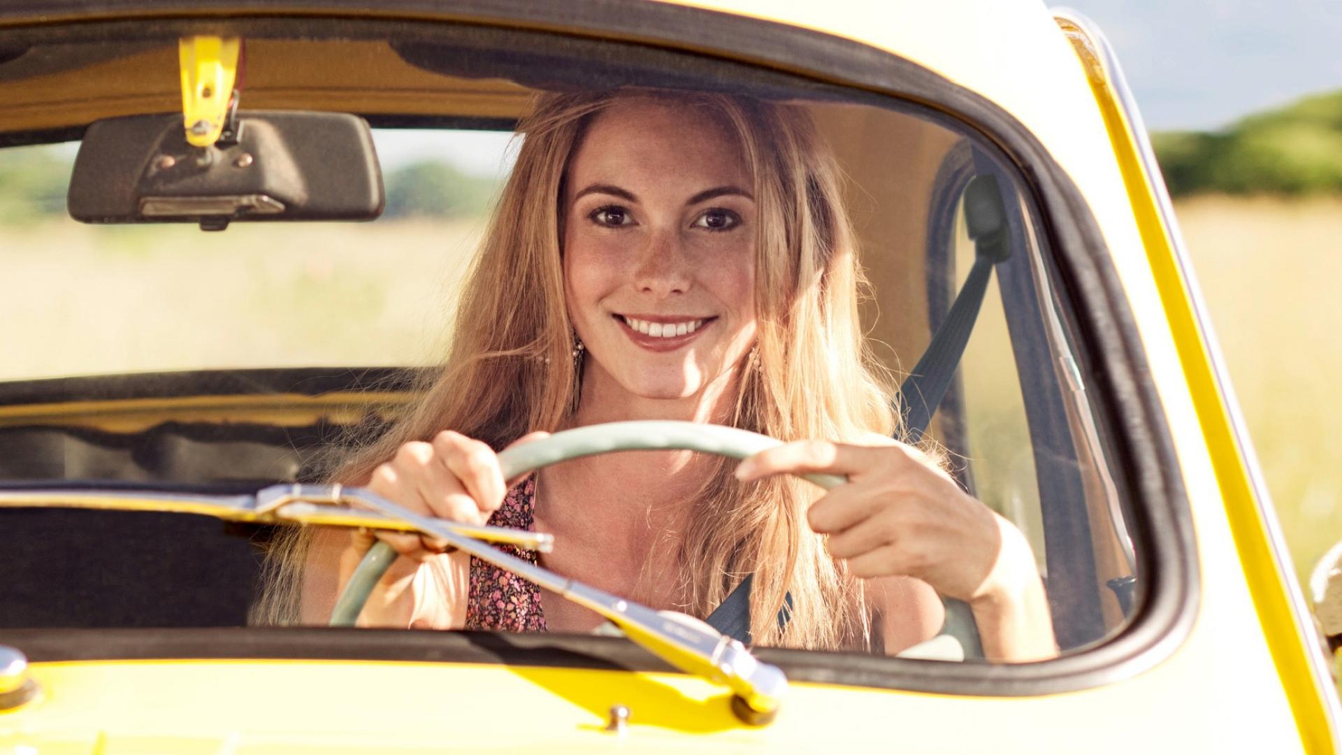 Eine Frau sitzt hinter dem Steuer eines gelben Autos und lächelt.