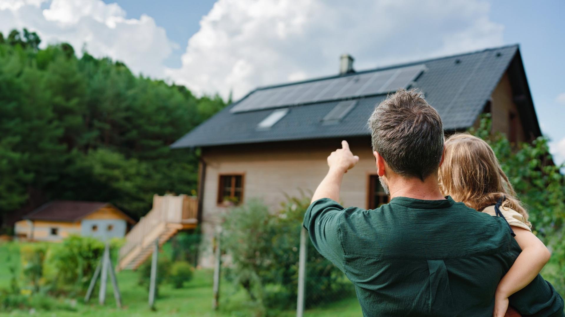 Ein Mann hält ein Kind auf dem Arm und zeigt auf ein Haus mit einer Photovoltaikanlage.