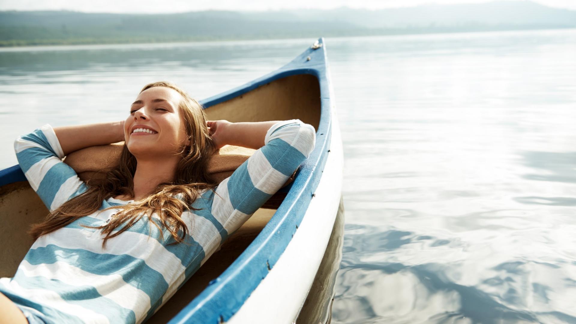 Eine glückliche, junge Frau liegt entspannt in einem kleinen Boot, das auf einem See schwimmt.