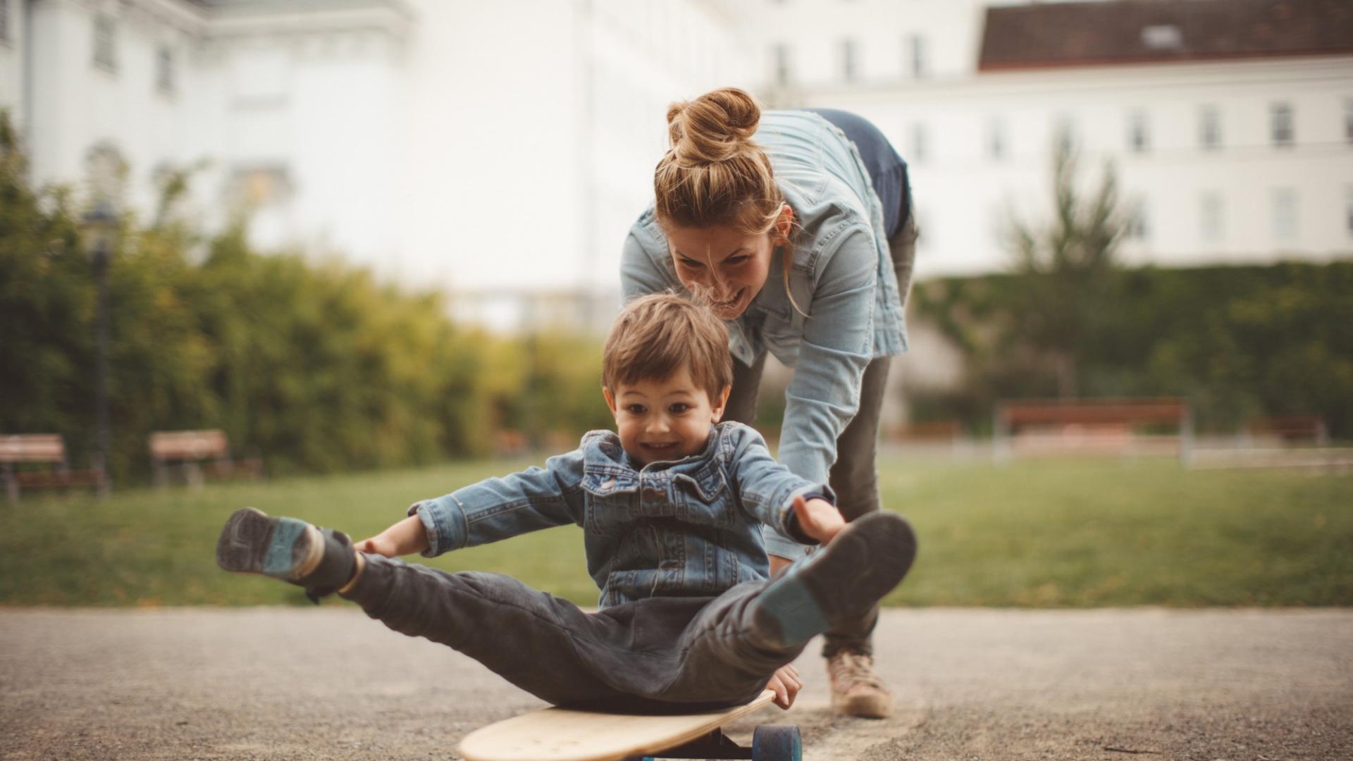 Eine Frau spielt mit einem Kind, das auf einem Skateboard sitzt, und schiebt es an.