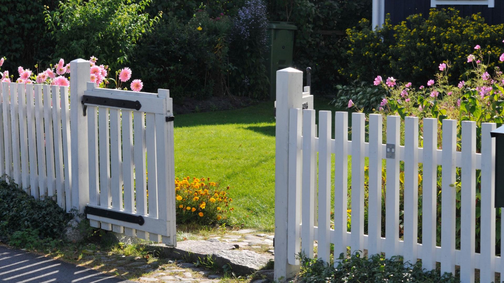 Ein offener weißer Gartenzaun vor einem Haus mit Wiese wird gezeigt