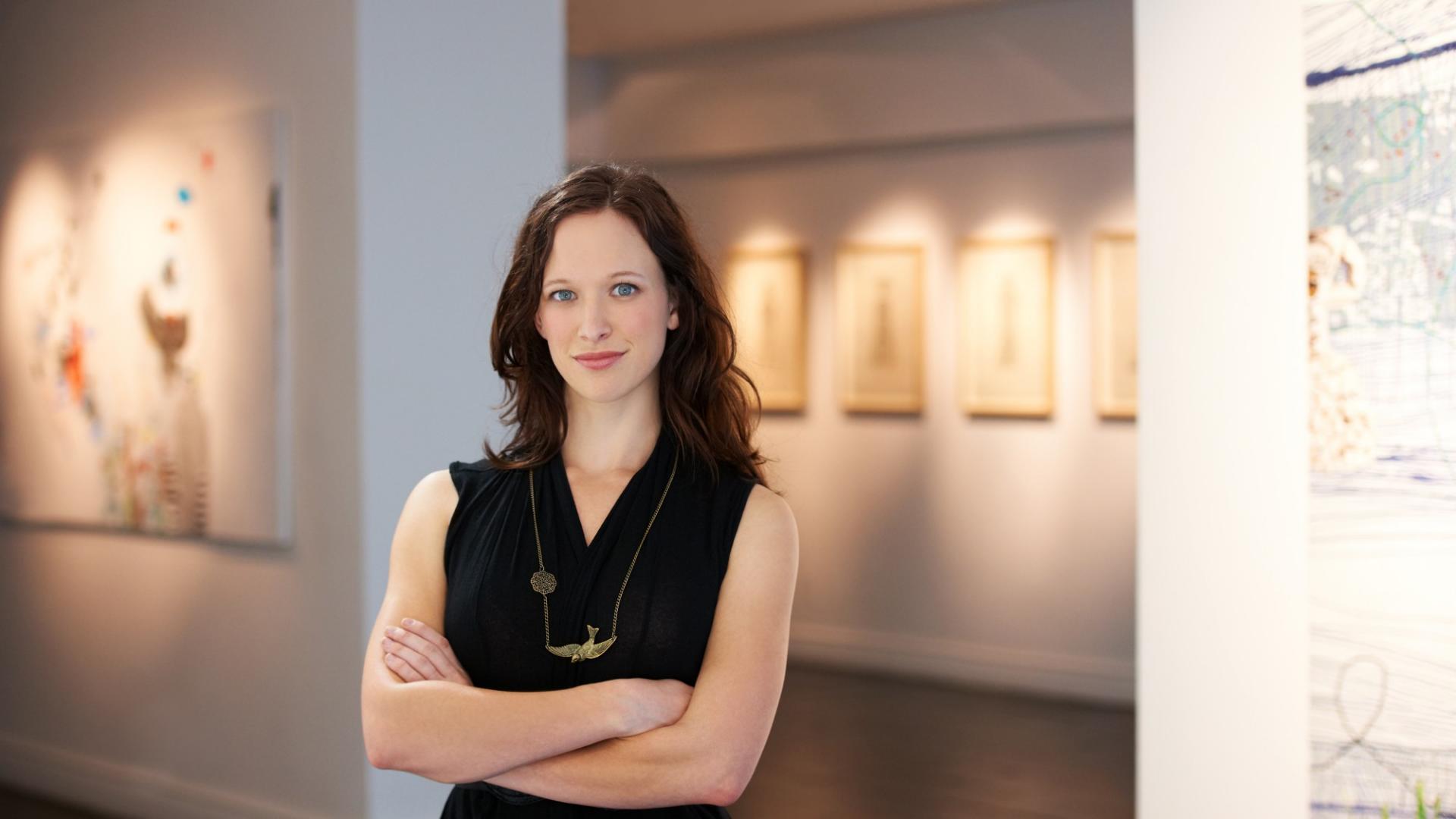 Eine junge Frau steht in einer Galerie und im Hintergrund sind Kunstgemälde zu sehen.