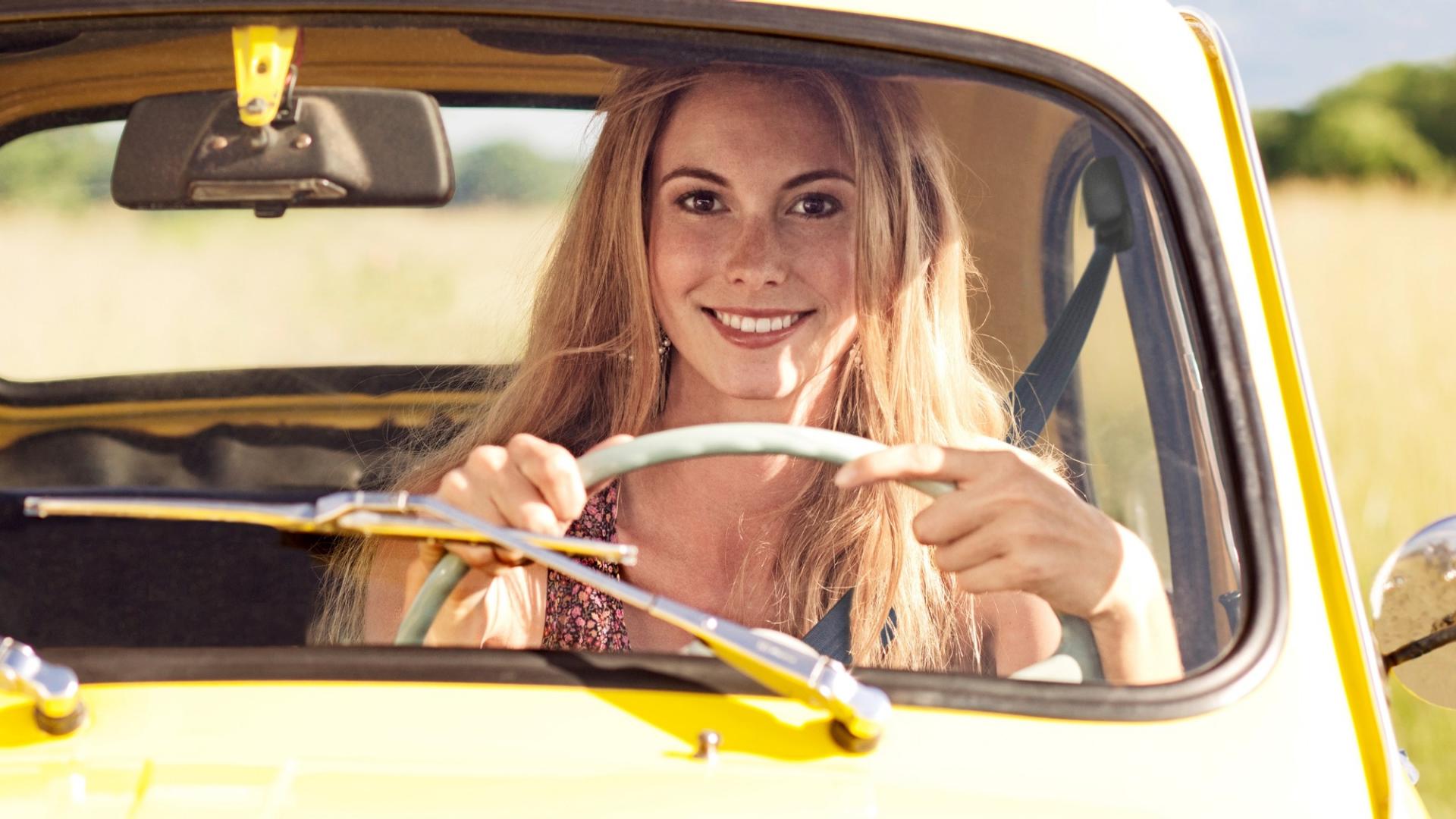 Eine blonde Frau fährt lächelnd ein gelbes Auto.