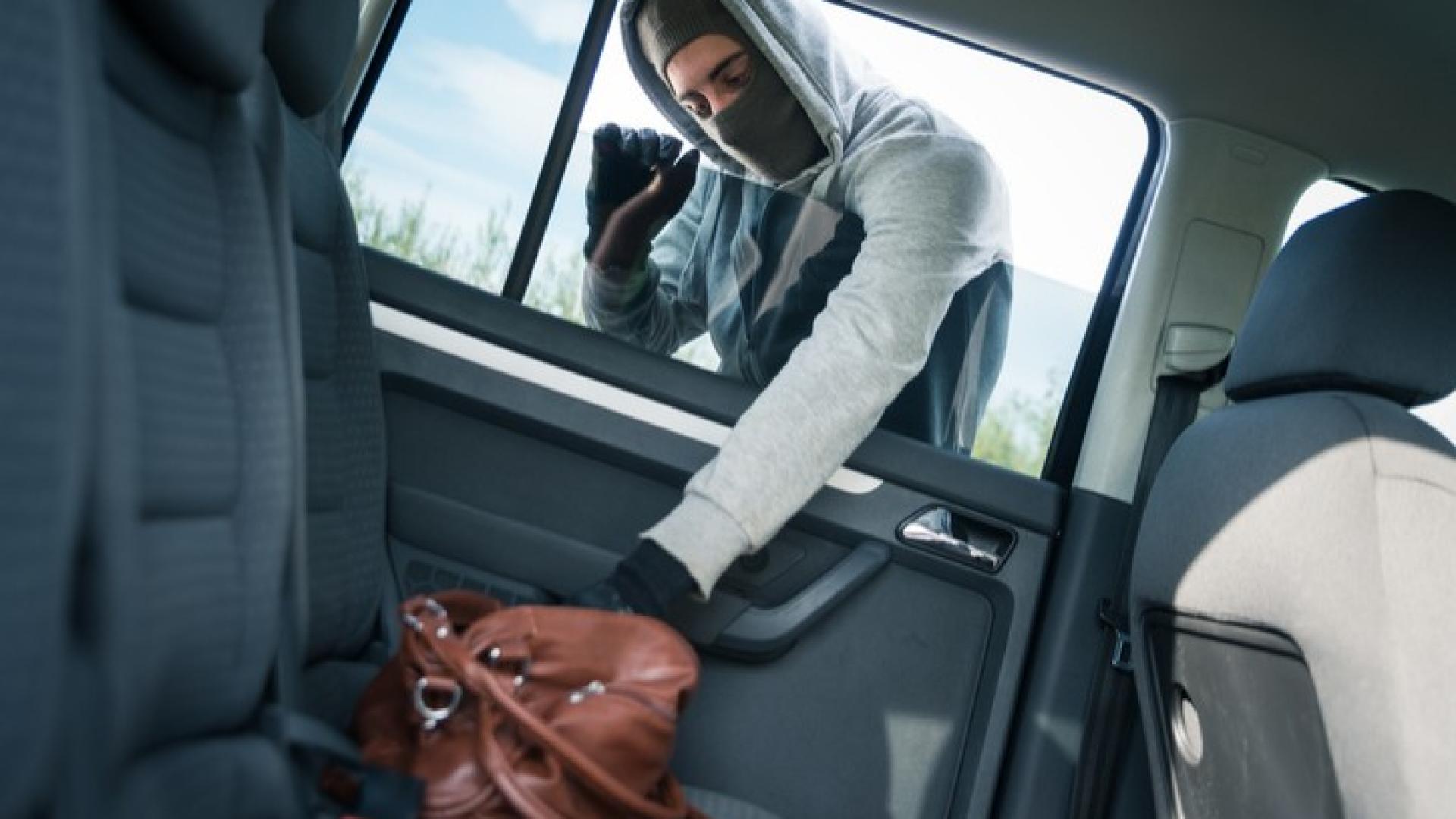 Ein maskierter Mann greift durch eine geöffnete Autofensterscheibe in ein Auto und wühlt in einer Tasche rum.