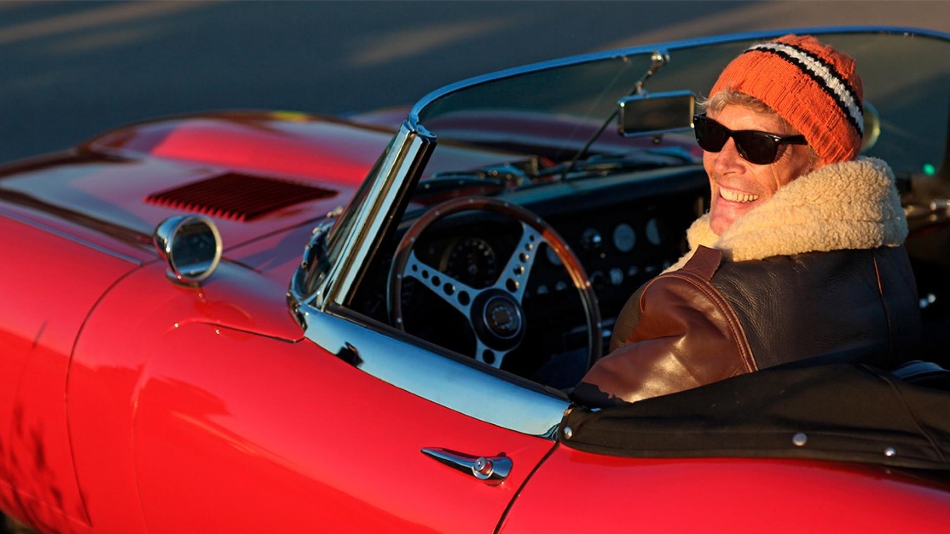 Ein Mann sitzt in seinem Cabrio-Oldtimer und lächelt mit Mütze auf dem Kopf in die Kamera