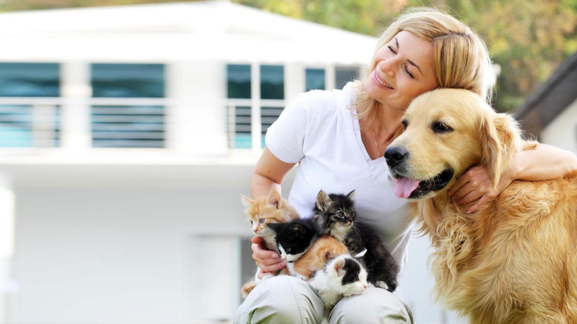 Eine blonde Frau kuschelt vor einem Haus mit ihren Tieren.