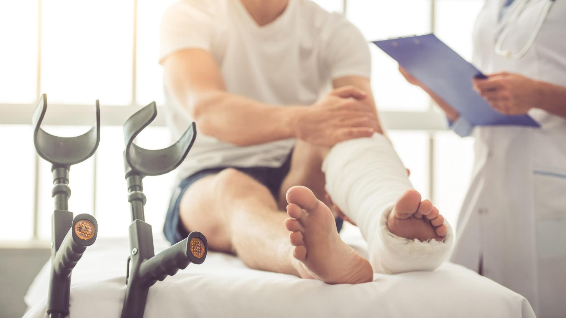 Ein Mann sitzt mit einem gebrochenen Fuß auf einem Krankenbett und wird von einem Arzt beraten. Neben ihm stehen die Krücken