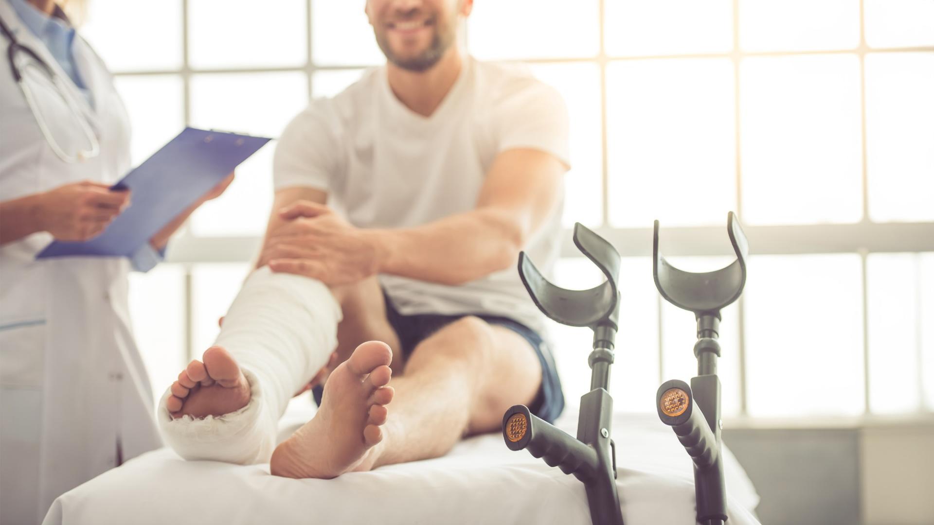 Ein Mann sitzt mit einem gebrochenen Fuß auf einem Krankenbett und wird von einem Arzt beraten. Neben ihm stehen die Krücken