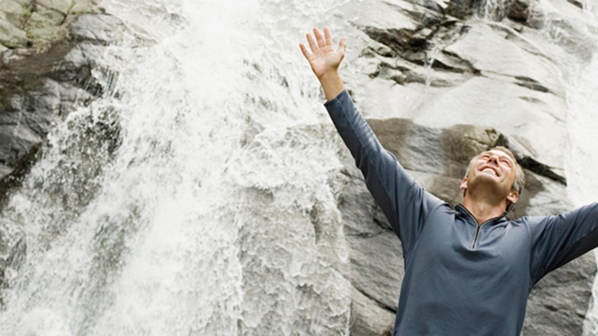 ein erwachsener Mann steht vor einer Felswand mit einem Wasserfall und streckt die Arme nach oben.