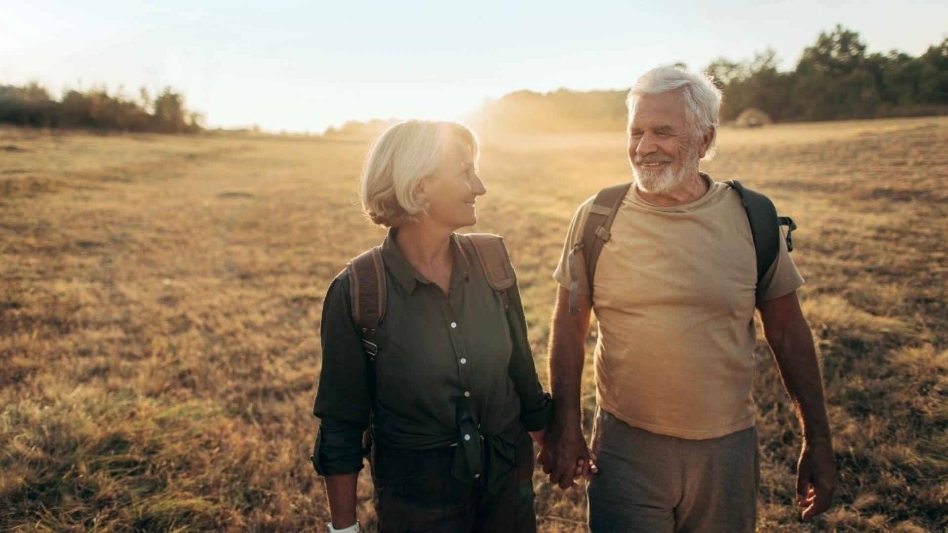 Ein Mann mit grauen Haaren und Bard und eine Frau mit grauen Haaren laufen Hand in Hand lächelnd über eine Wiese.