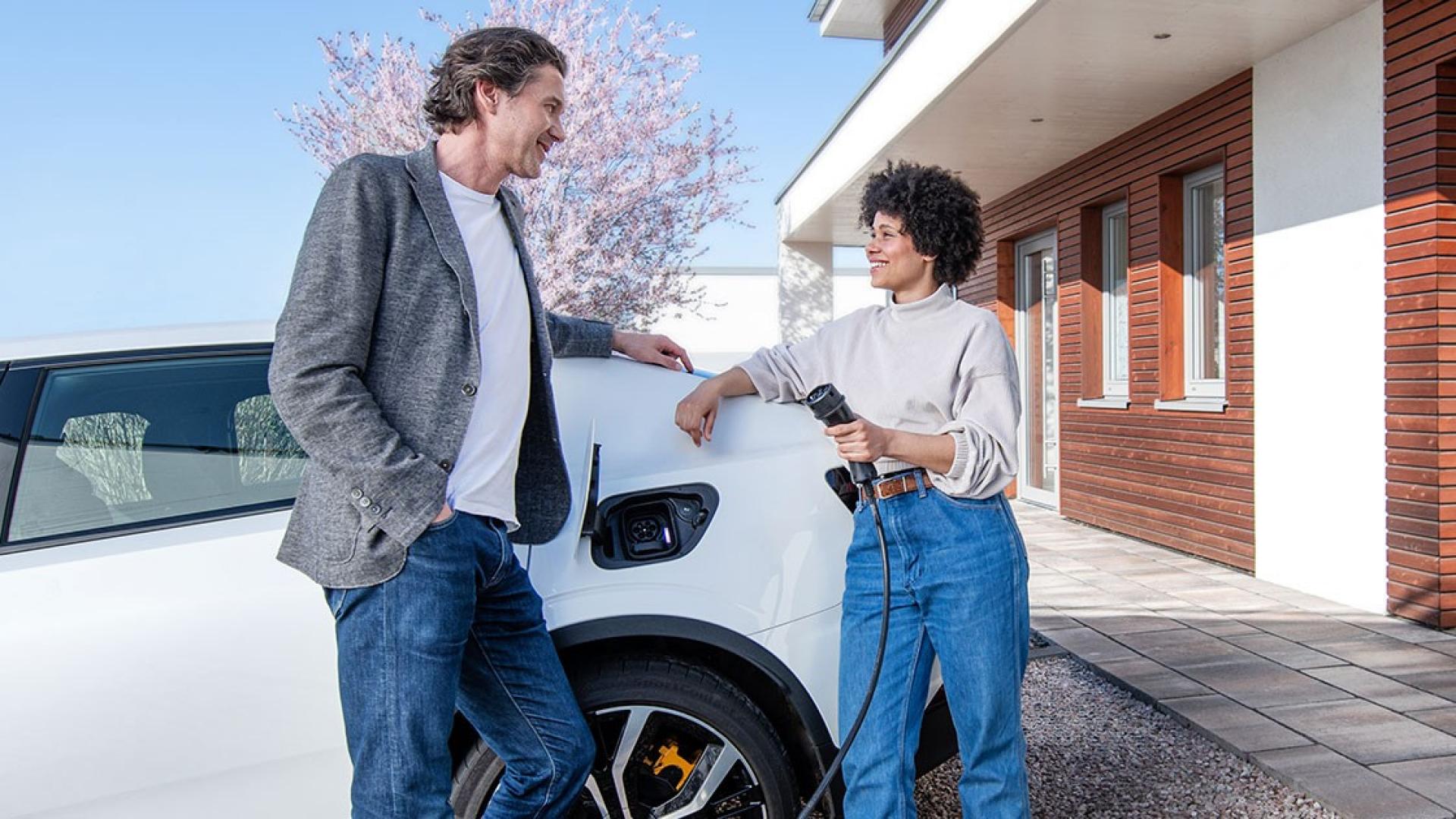 Ein Mann und eine Frau, welche ein Ladekabel in der Hand hält, lehnen sich an ein Elektroauto.