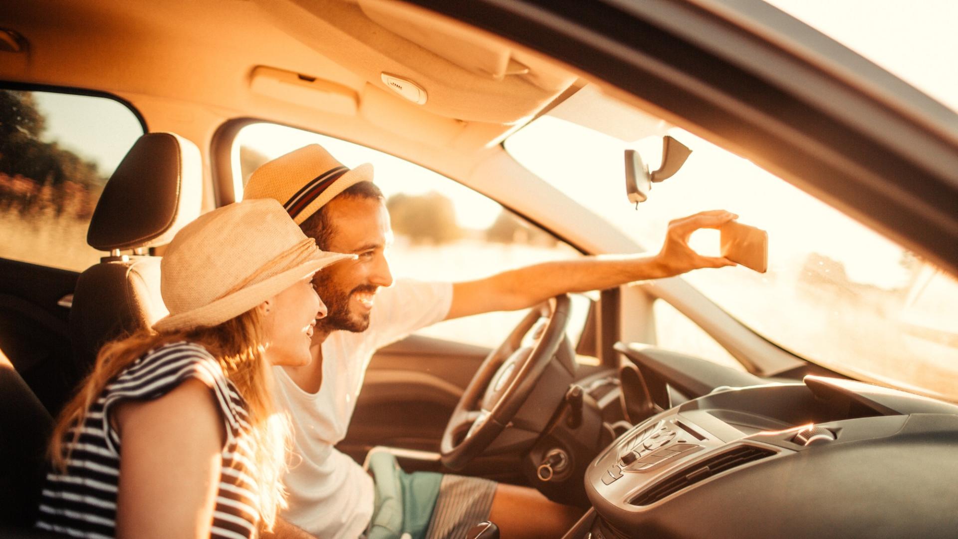 Eine Frau und ein Mann mit Hut sitzen lächelnd in einem Auto und machen ein Selfie.