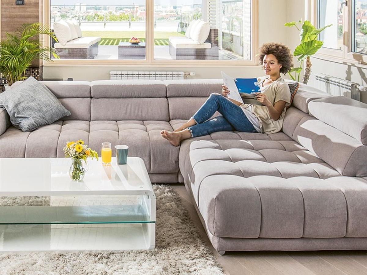 Eine junge Frau sitzt entspannt auf einer großen Couch in einem Wohnzimmer und ließt ein Prospekt.