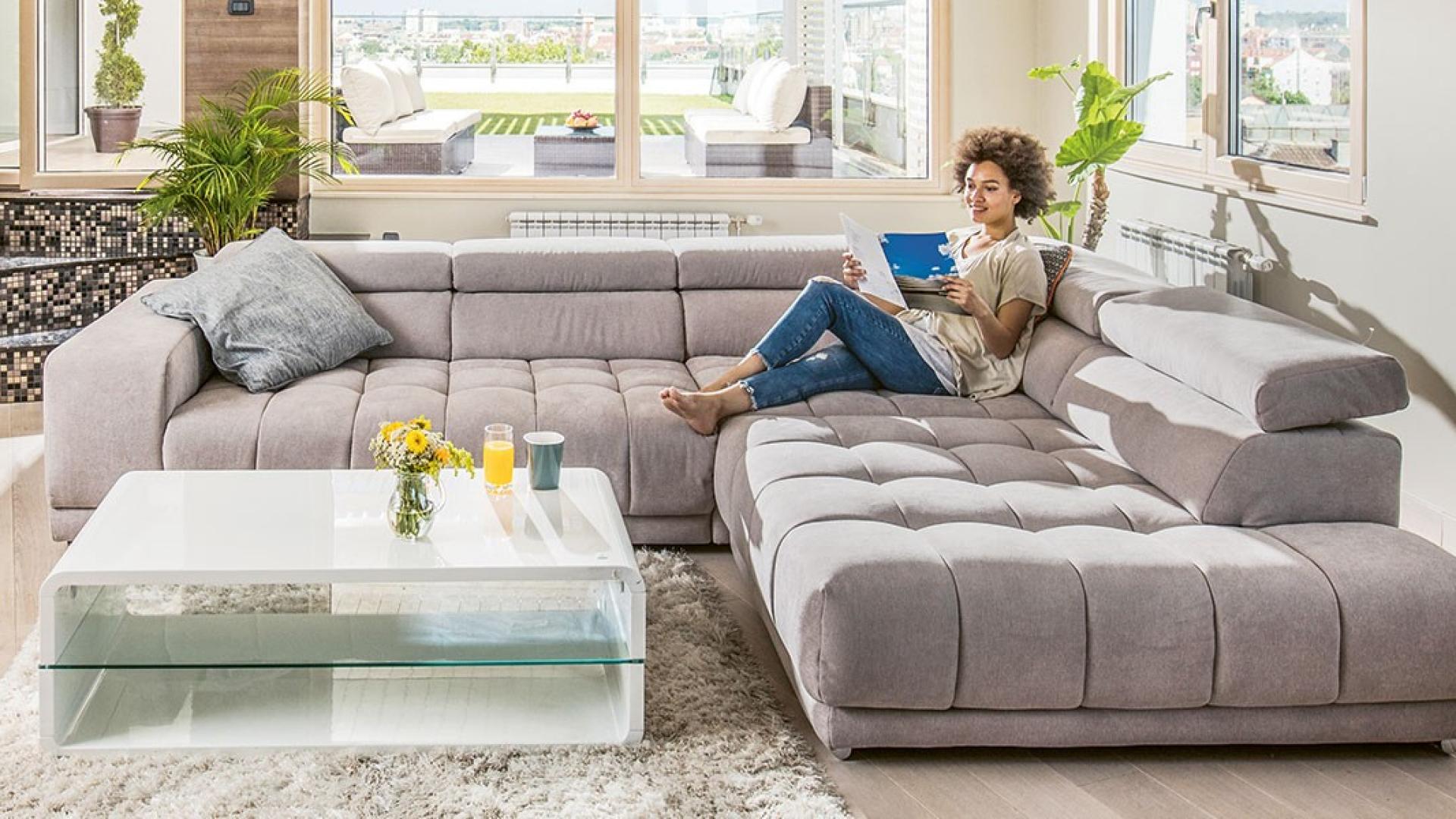 Eine junge Frau sitzt entspannt auf einer großen Couch in einem Wohnzimmer und ließt ein Prospekt.