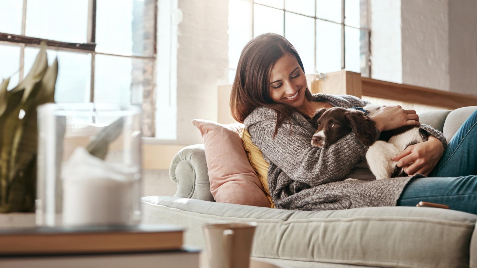 Auf dem Bild ist eine junge Frau auf ihrer Couch zu sehen, die mit ihrem Hund kuschelt.