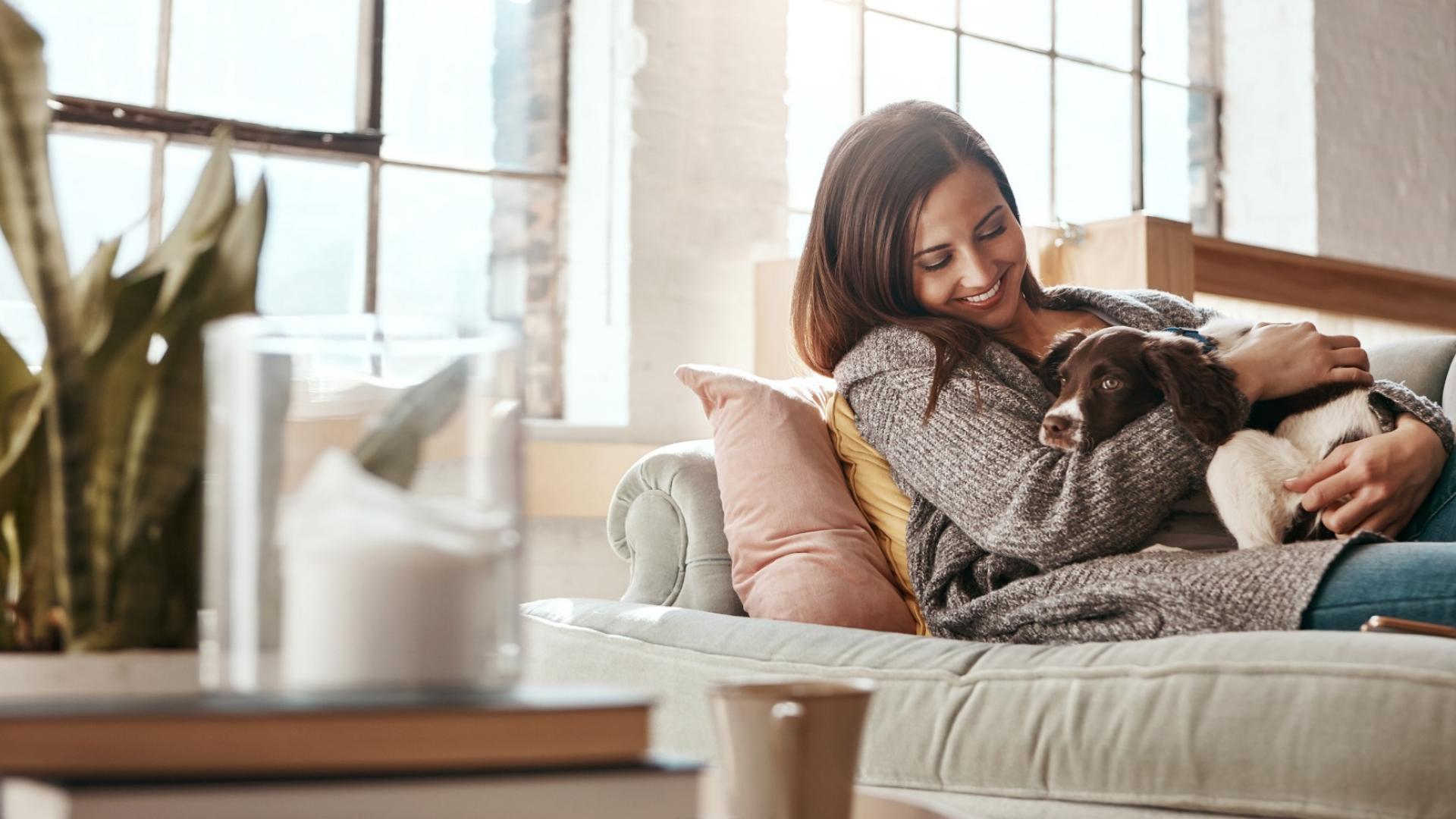 Auf dem Bild ist eine junge Frau zu sehen, die auf ihrer Couch mit ihrem Hund kuschelt.