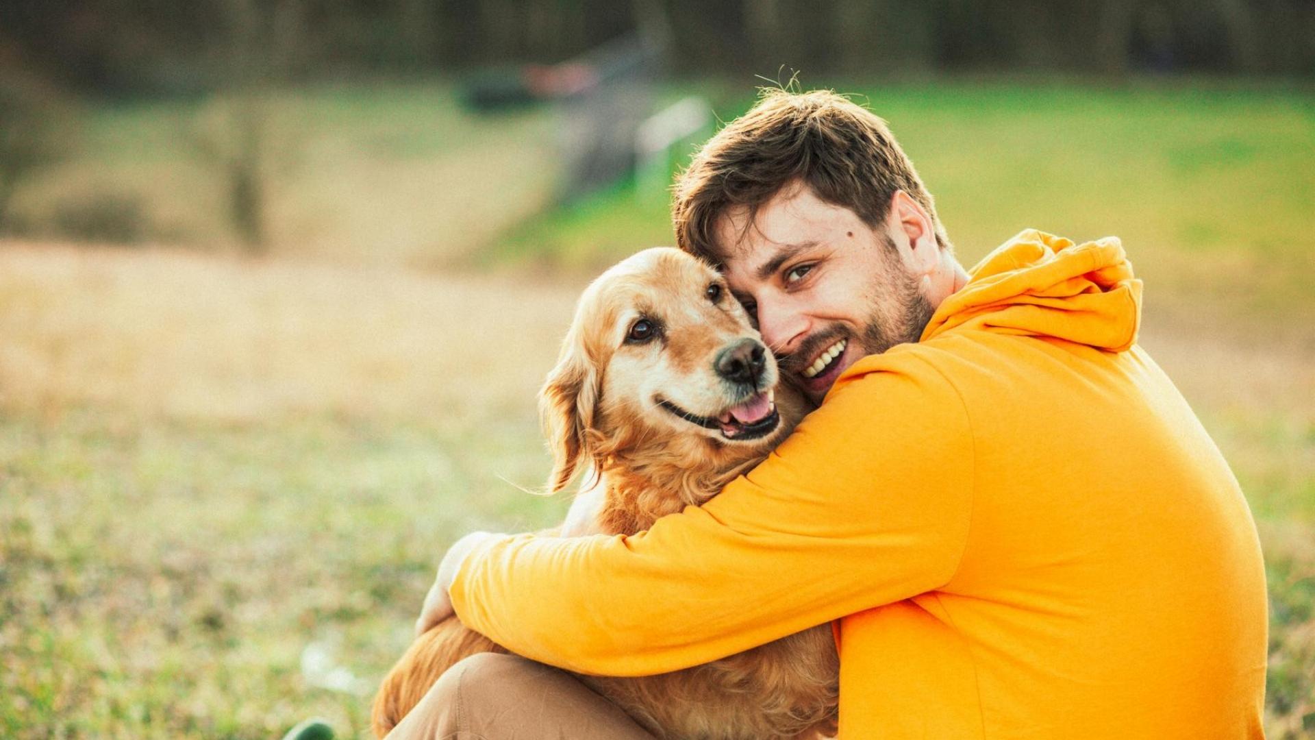 Ein junger Mann sitzt auf einer Wiese und umarmt lächelnd einen Hund.