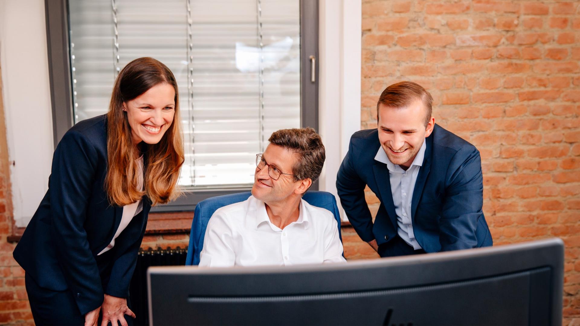 Eine Frau und zwei Männer in Geschäftskleidung blicken auf einen Monitor und lächeln